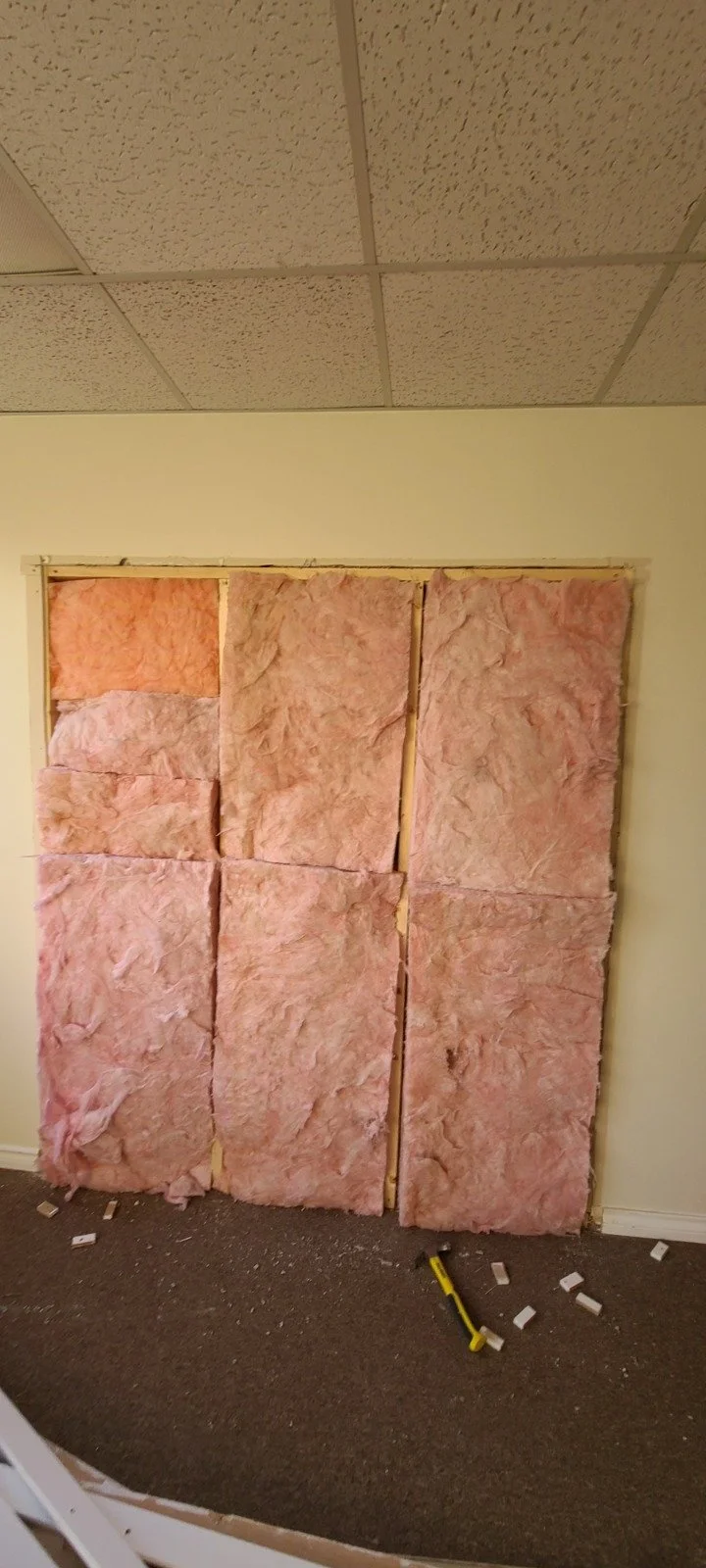 Interior wall under construction with pink insulation batts installed between wooden studs. Debris and a hammer are on the carpeted floor.