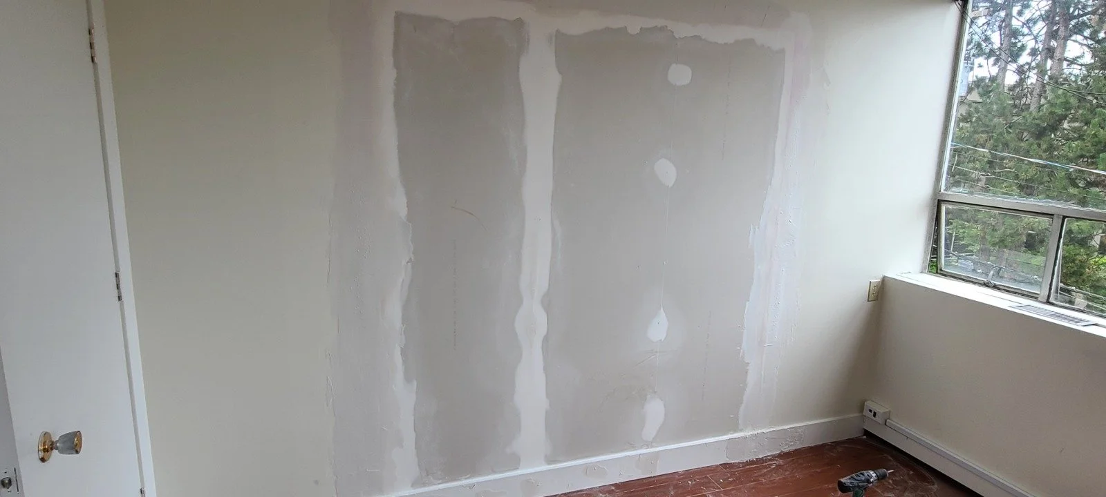 Interior room wall with drywall in progress, partially patched, next to a window with a view of trees outside.