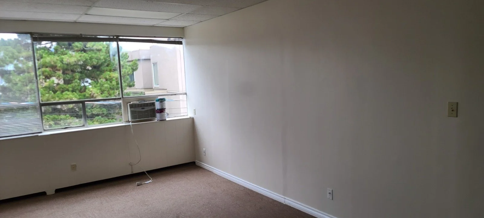 Empty room with large window, air conditioning unit, and beige carpet.