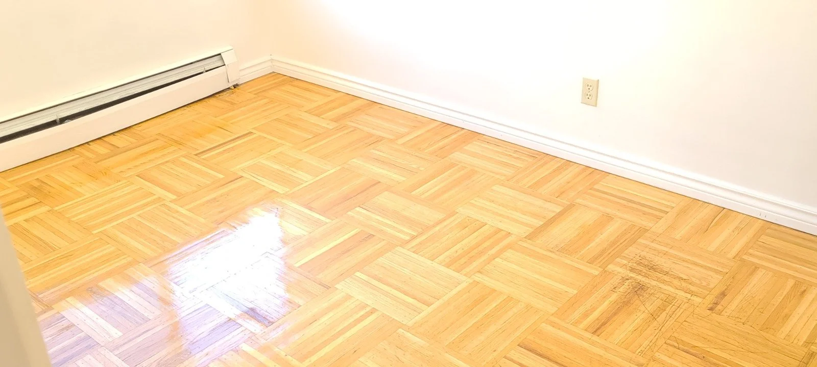 Empty room with hardwood floor, white walls, white baseboard, a black and white baseboard heater, and a wall electrical outlet.