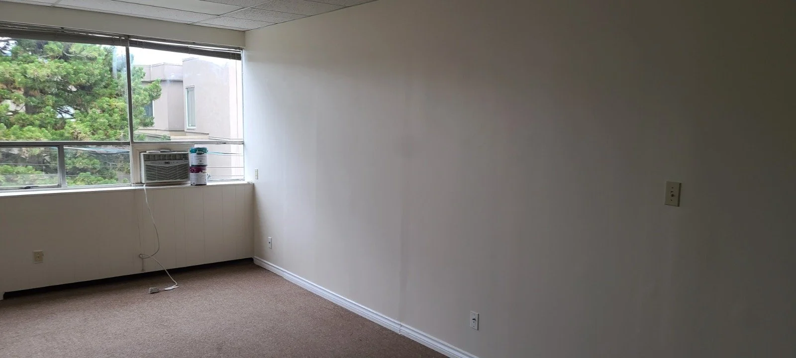 Empty room with beige carpet, white walls, large window showing greenery outside, window air conditioning unit and can of paint on sill, electrical outlets on walls.