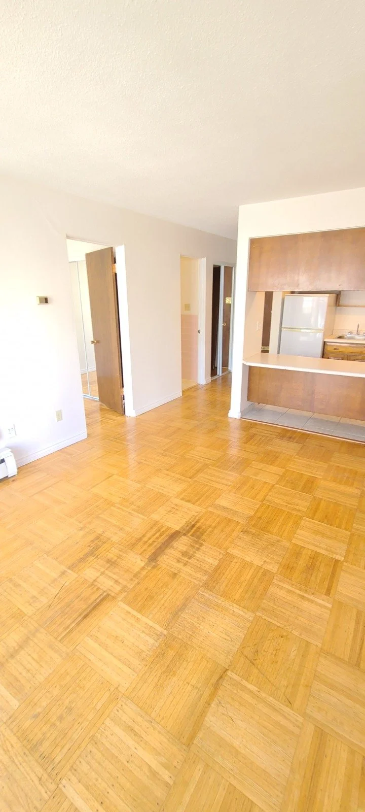An empty room with wooden parquet flooring and white walls, leading to a kitchen area with a refrigerator and wooden cabinets.