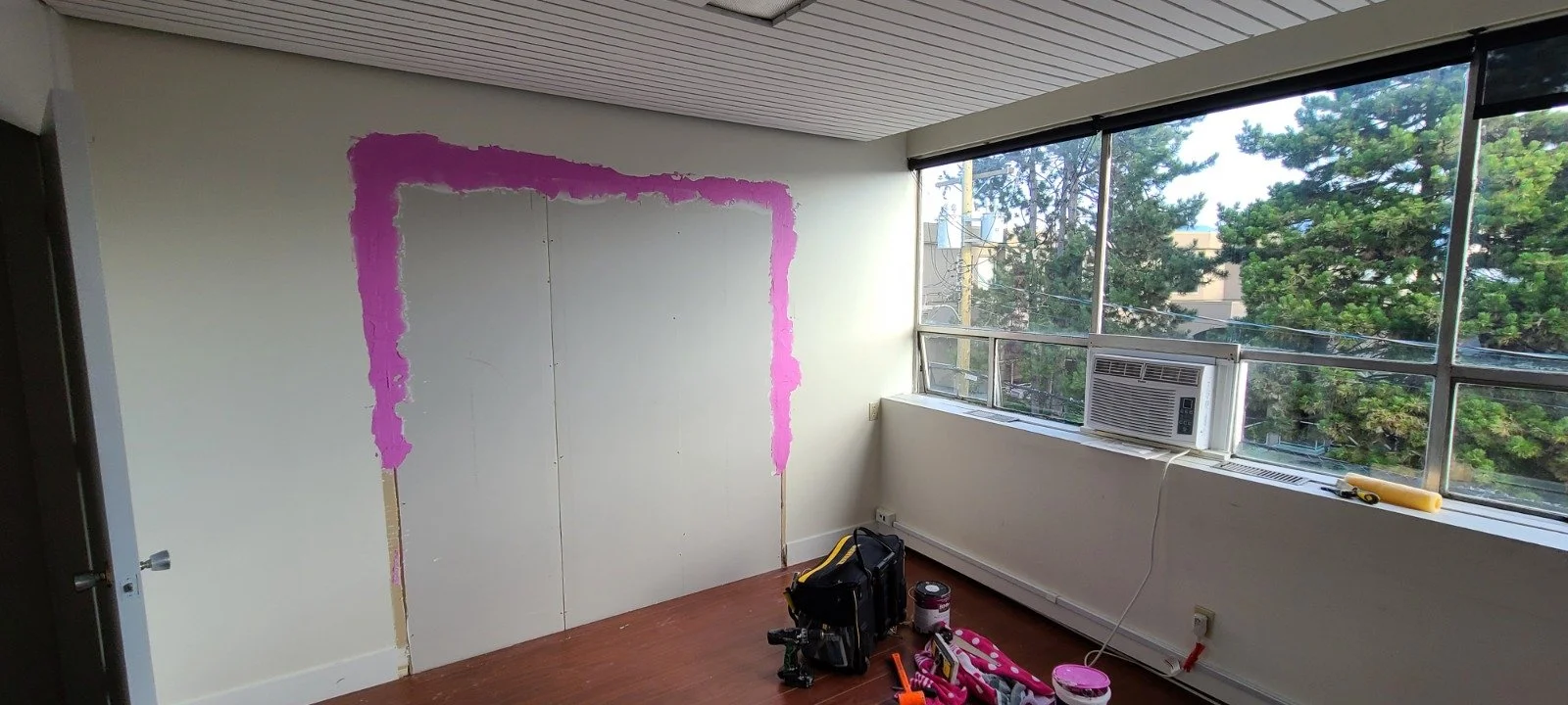 Room with a partially painted wall, pink paint around the edges, large windows with an air conditioning unit, and construction tools and supplies on the floor.