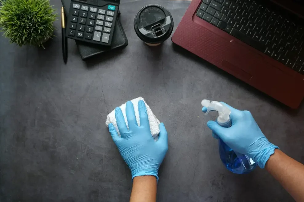 Person wearing blue gloves cleaning a dark desk with a white cloth and spray bottle. Office supplies like a calculator, a coffee cup, a laptop, a pen, and a small green plant are visible.