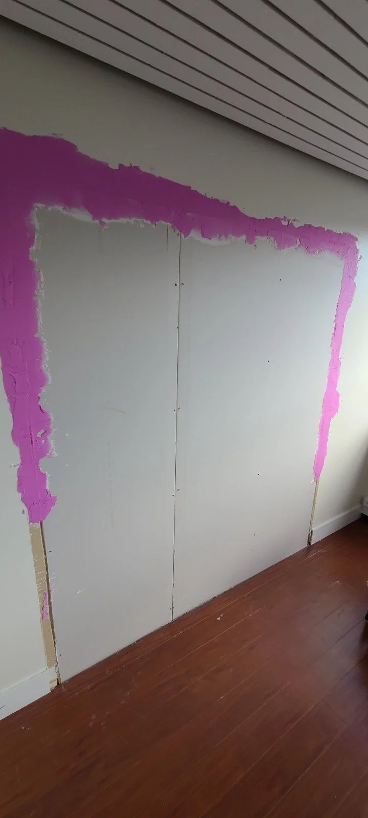 Partially painted wall with pink primer or paint around the edges in a room with a hardwood floor and a ceiling with horizontal panels.