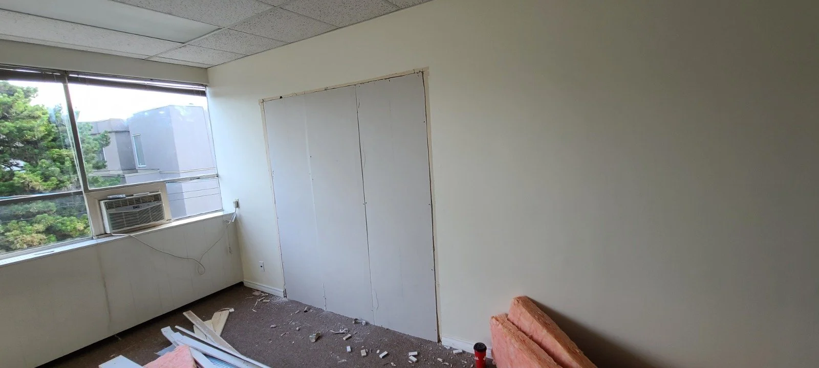 Empty room with a window air conditioning unit, debris on the floor, and a partially constructed wall or doorway.