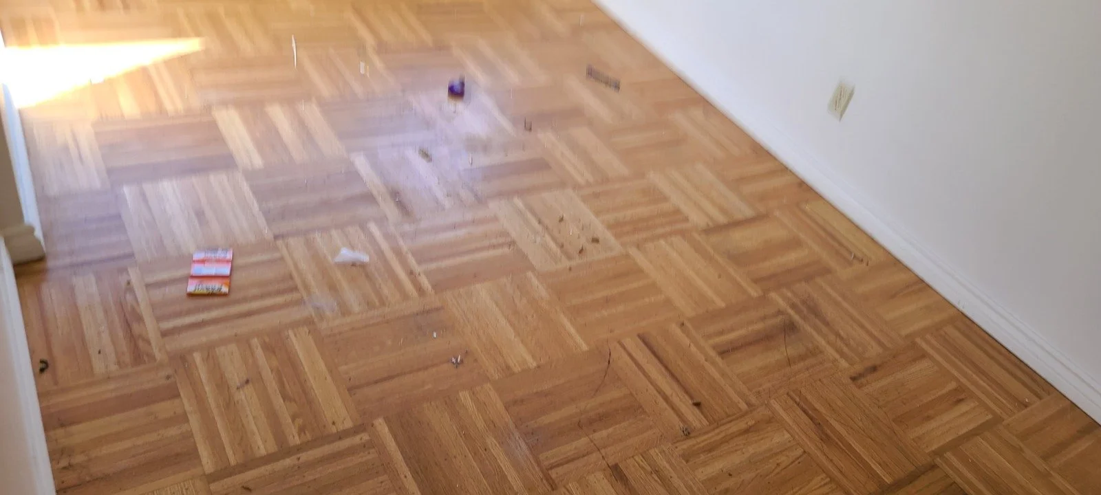 A hardwood floor with scattered debris and a small colorful object near a wall with wall outlet, sunlight shining on the floor.