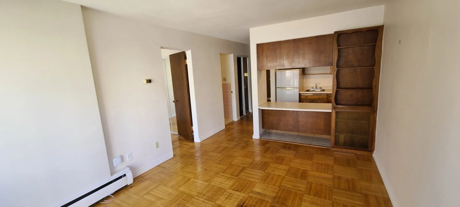 Empty living room with hardwood floors, white walls, a small open kitchen with a refrigerator, and built-in wooden shelves on the right side.