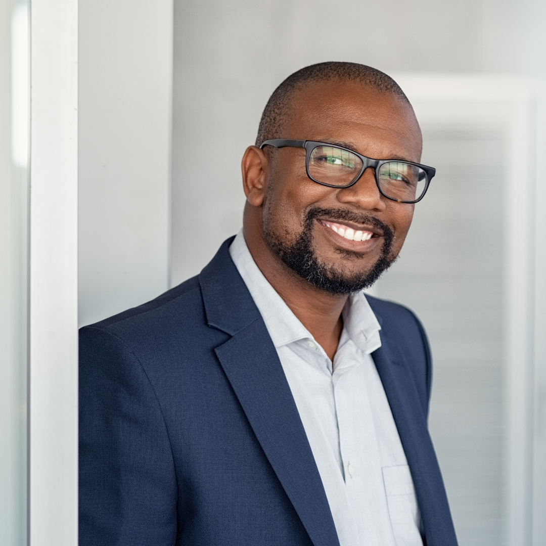 Professionally dressed, smiling black man with glasses leaning against a wall