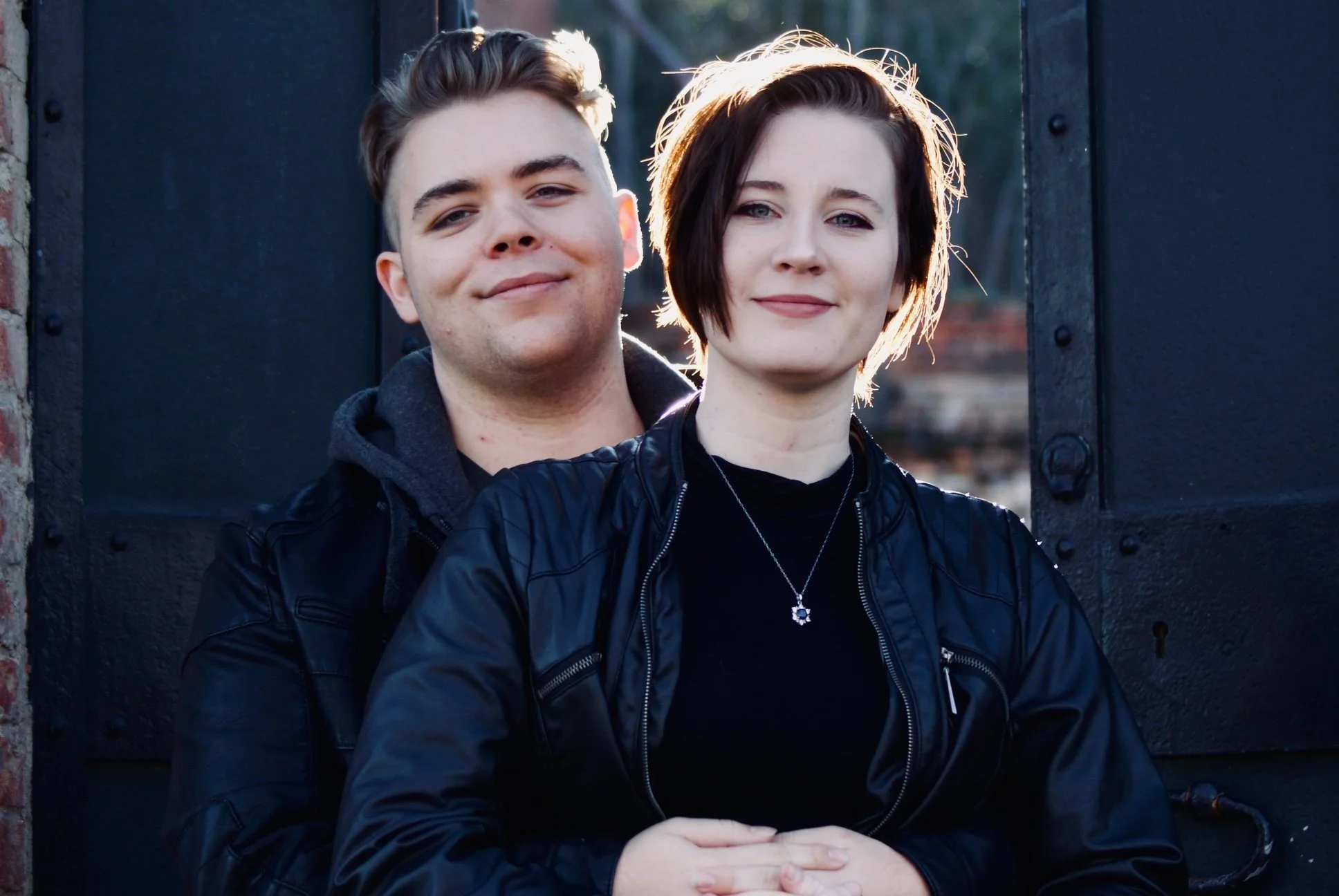 A young couple smiling, looking in the camera while leaning against eachother.