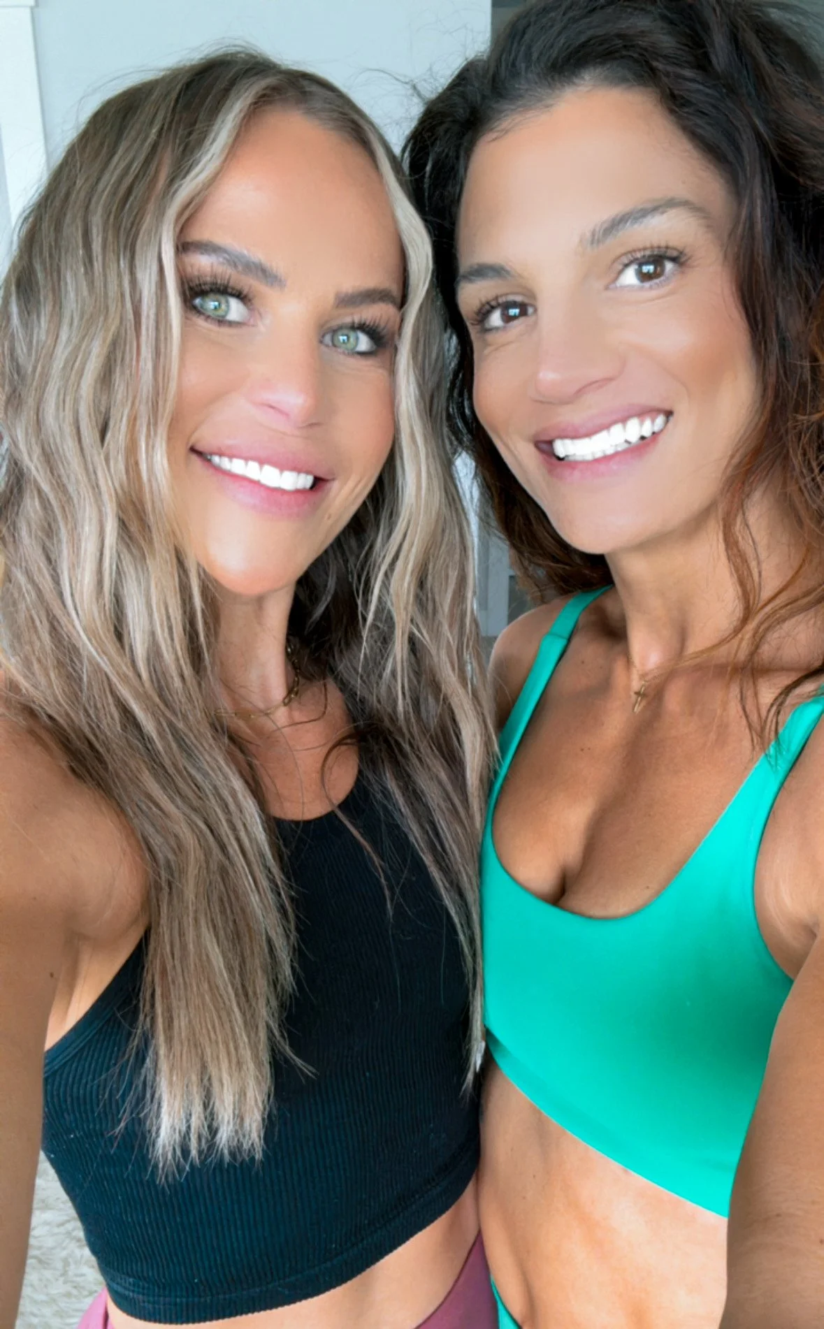 Two women smiling, wearing tank tops, taking a selfie indoors.