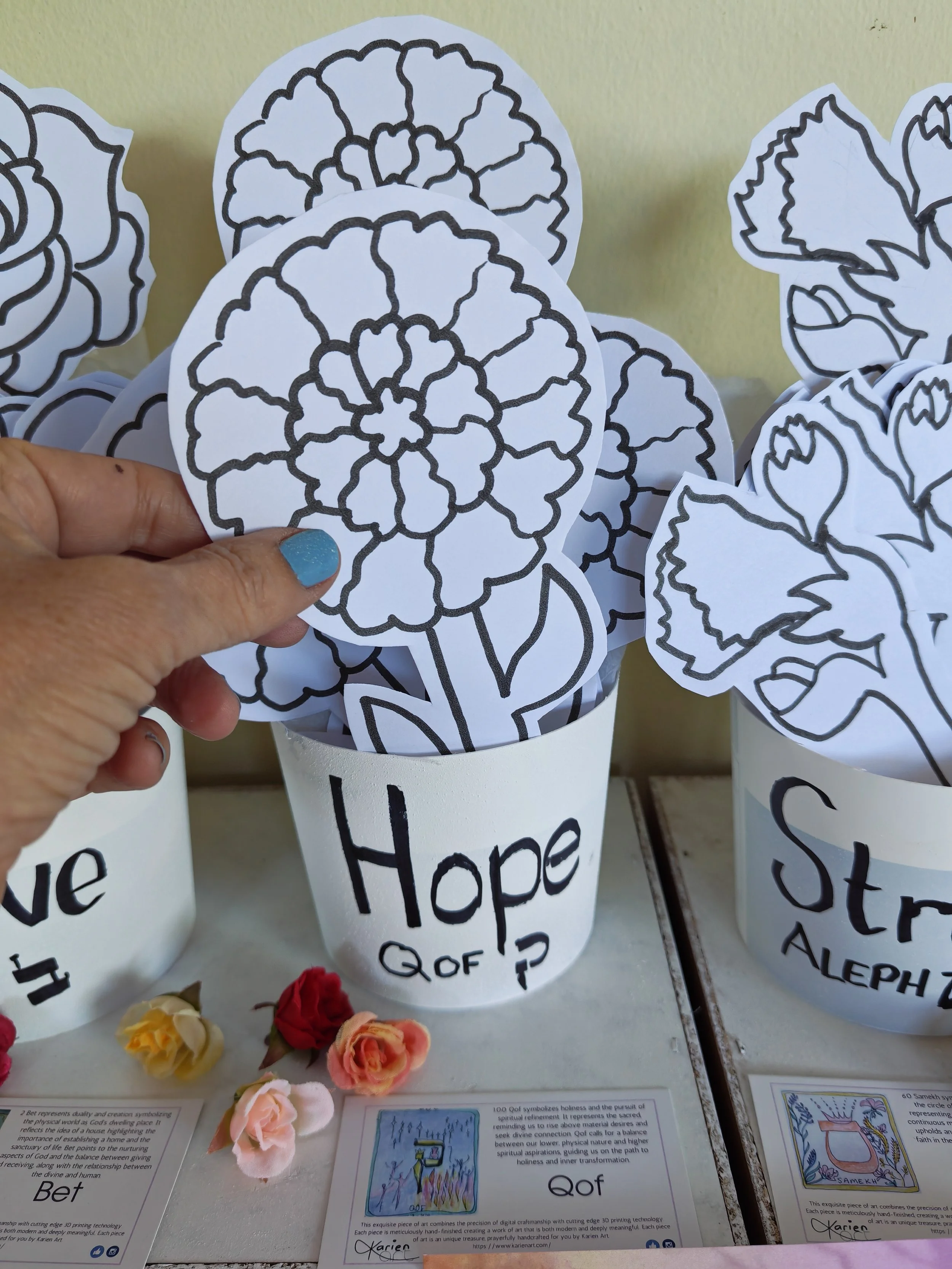 Paper cutouts of flowers with black outlines, labeled with words such as 'Hope' and 'Bet,' arranged on display alongside paper flowers and informational sheets.