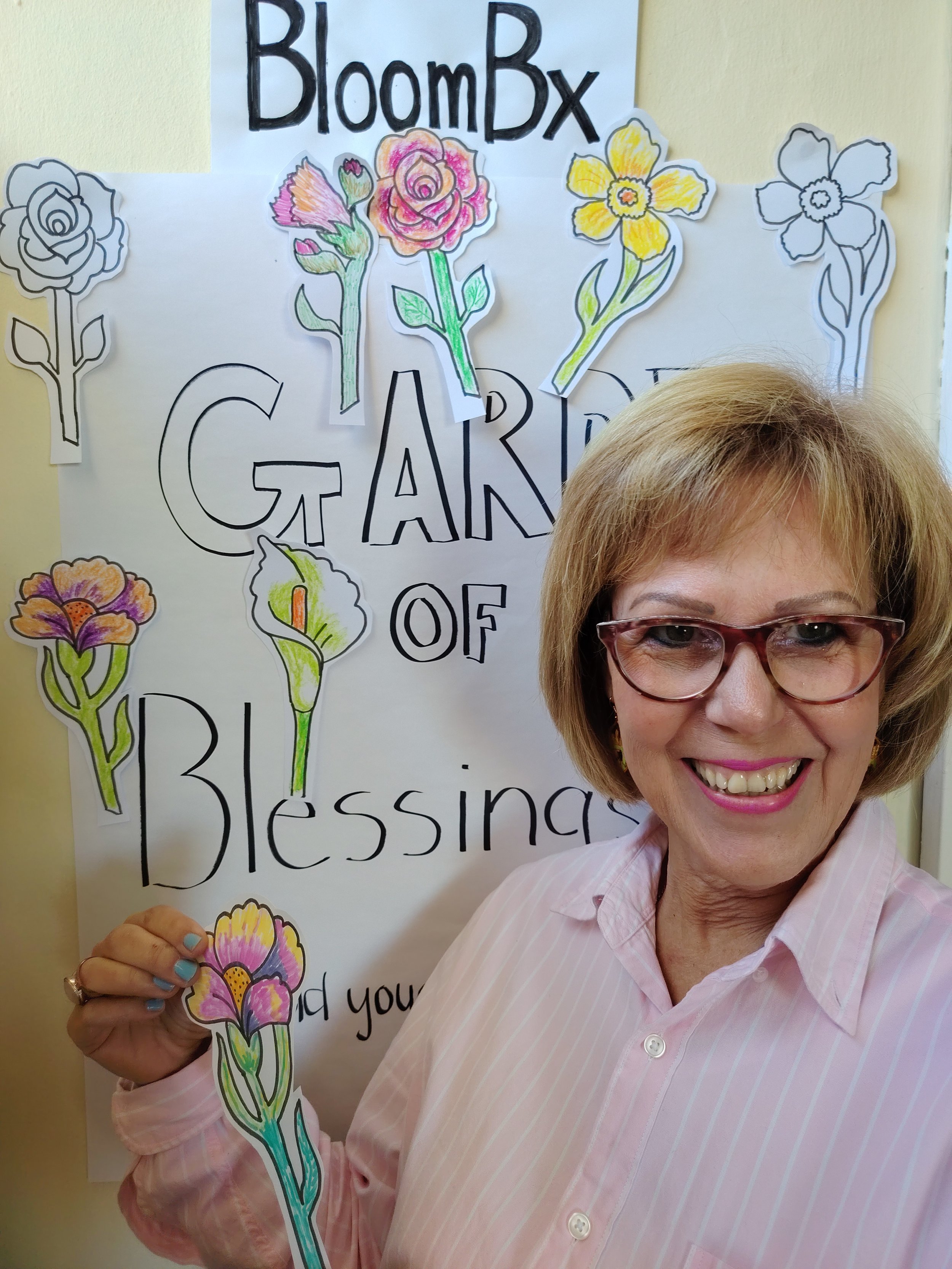 A smiling woman with short blonde hair and glasses standing in front of a decorated board. The board has the words 'BloomBox GARDEN of Blessings' written on it, with colored and outlined flowers attached to the background.