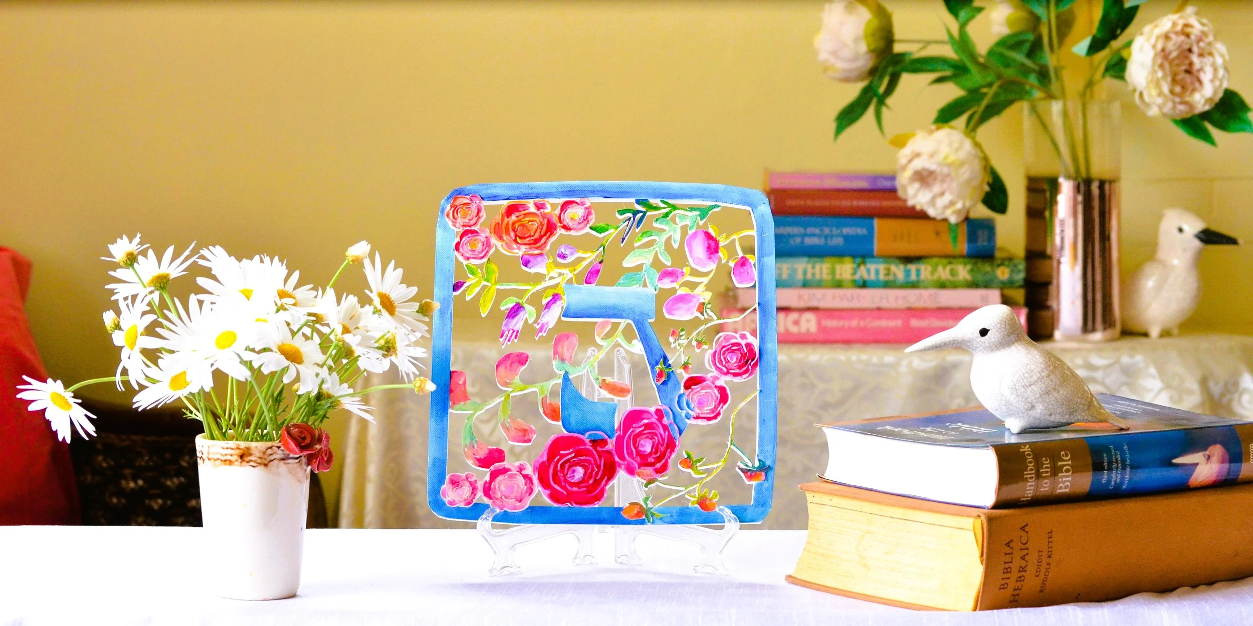 A decorative table with a bouquet of white daisies in a white and brown vase, a colorful floral wall art, a stack of books, and small ceramic bird figurines on top of the books and behind them, with a large plant with pink flowers in the background.