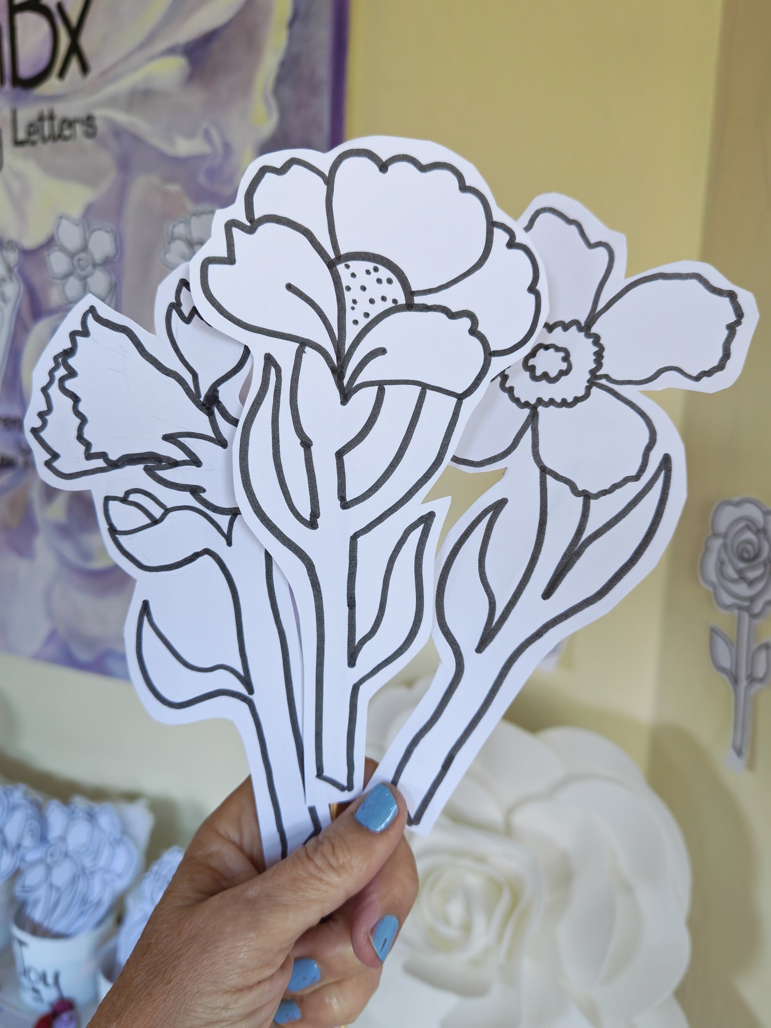 Black and white floral coloring sheets being held in a hand with blue nails. There are more sheets and paper flowers in the background.