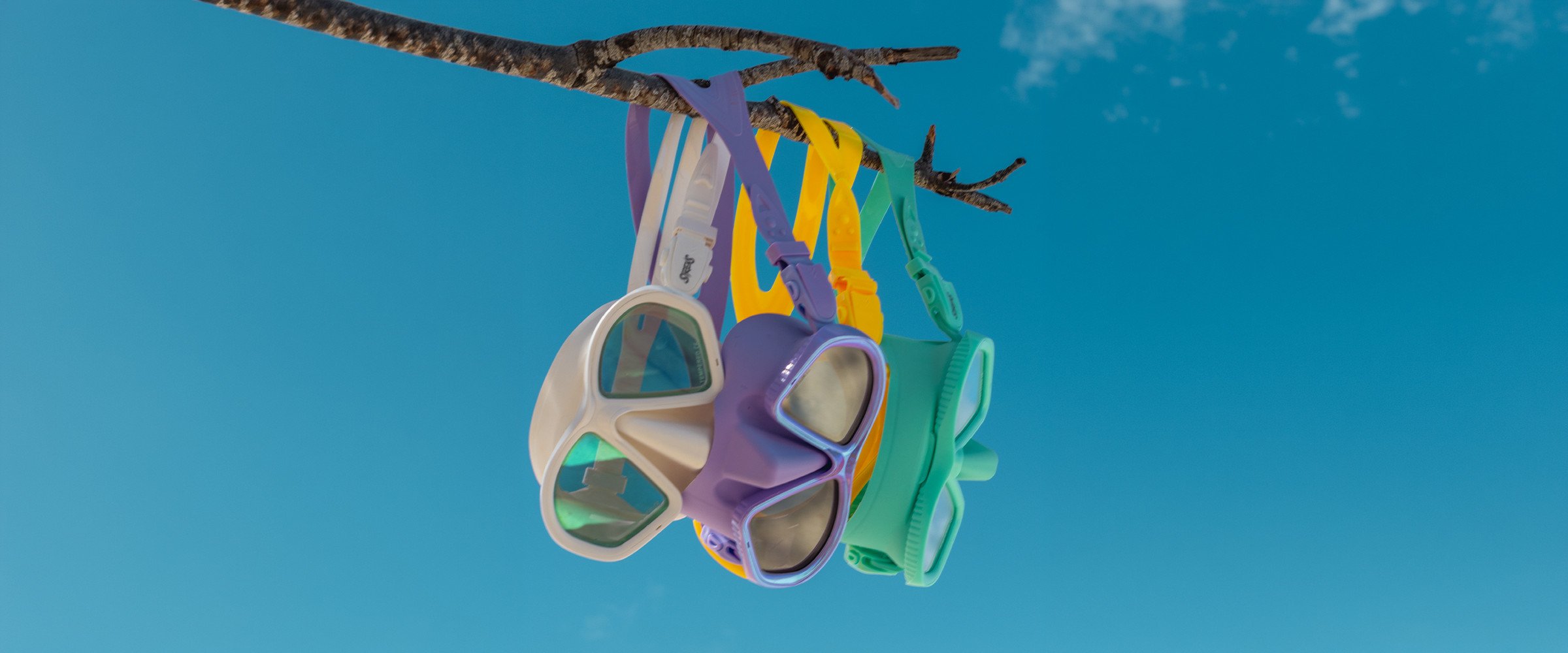 White, lavender, yellow and aqua Sirens dive masks hanging against a clear blue sky.