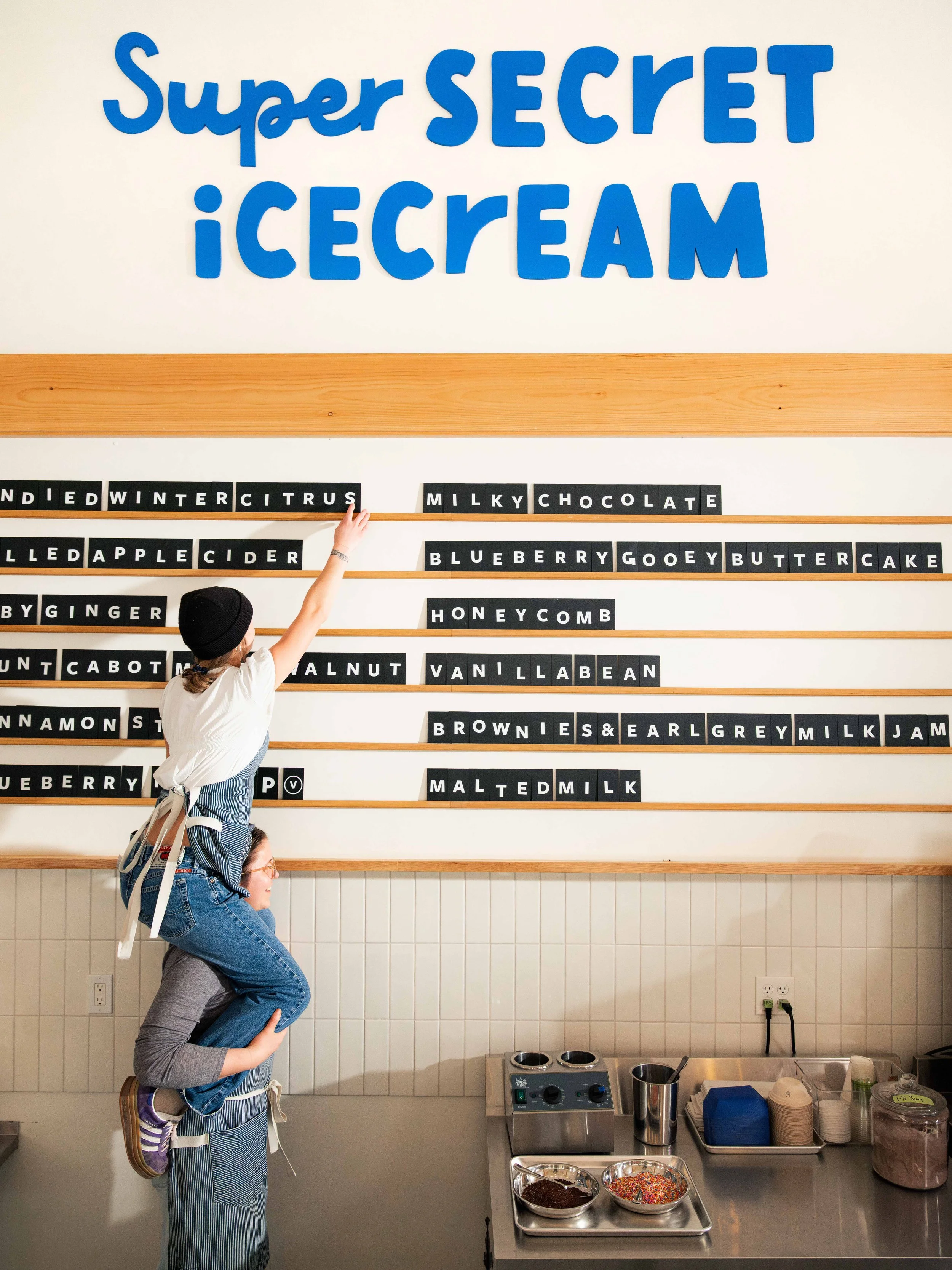 A person carrying another person on their shoulders in front of a wall with a sign that reads 'Super SECRET ICE CREAM' and a selection of toppings in bowls on a counter.