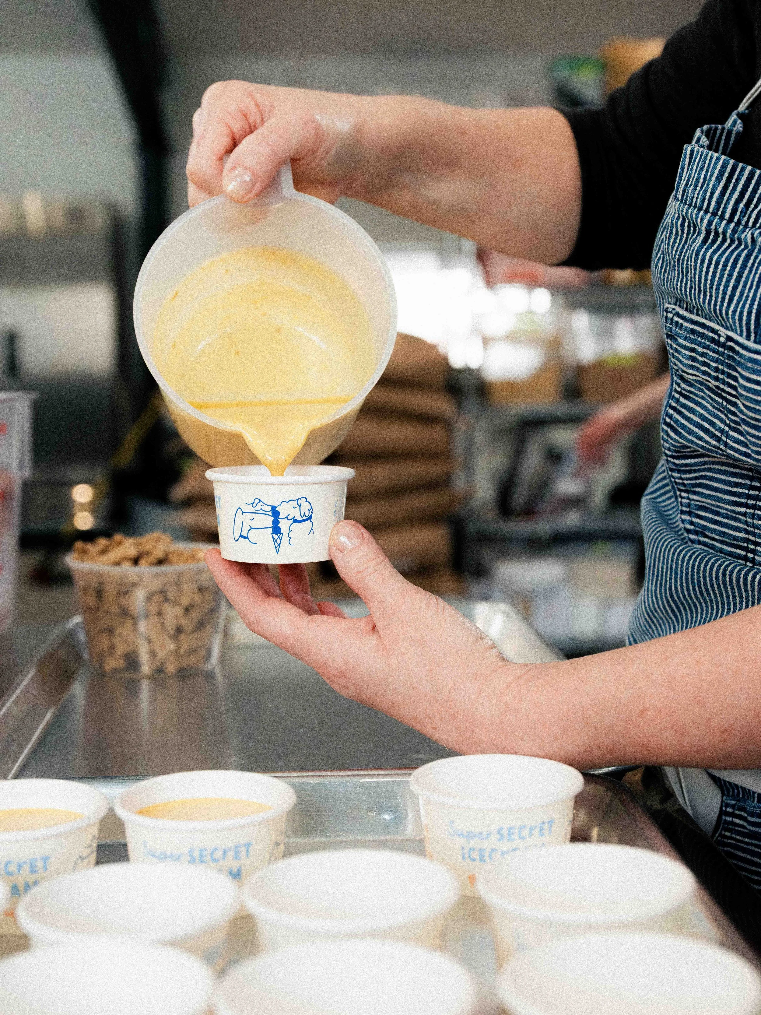 A person wearing a black shirt and striped apron pours yellow ice cream from a container into small white cups labeled 'Super SECRET ICE CREAM' on a stainless steel work surface.