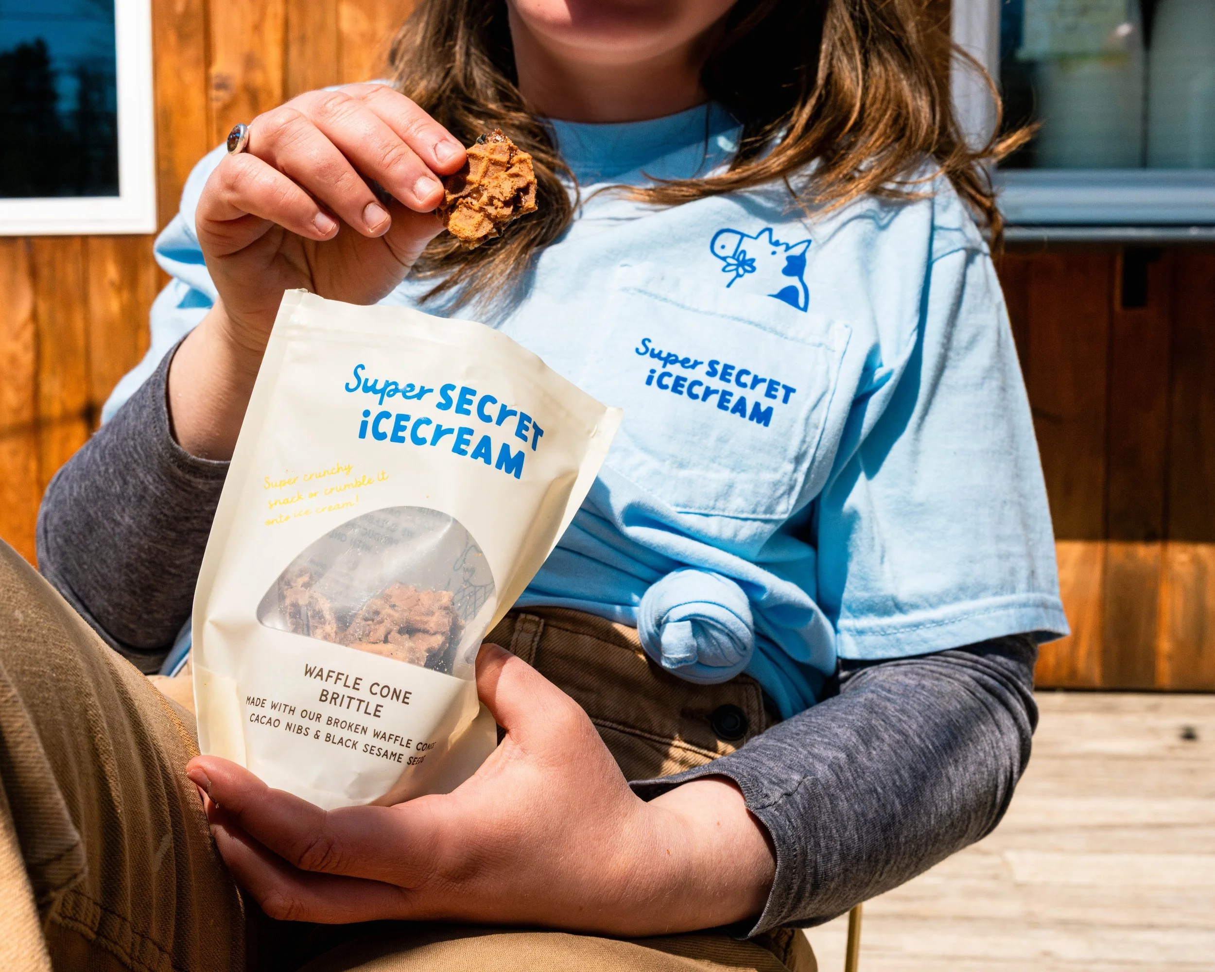 Person with brown hair wearing a light blue shirt holding a bag of waffle cone brittle ice cream with one hand, sitting outdoors against a wooden fence.