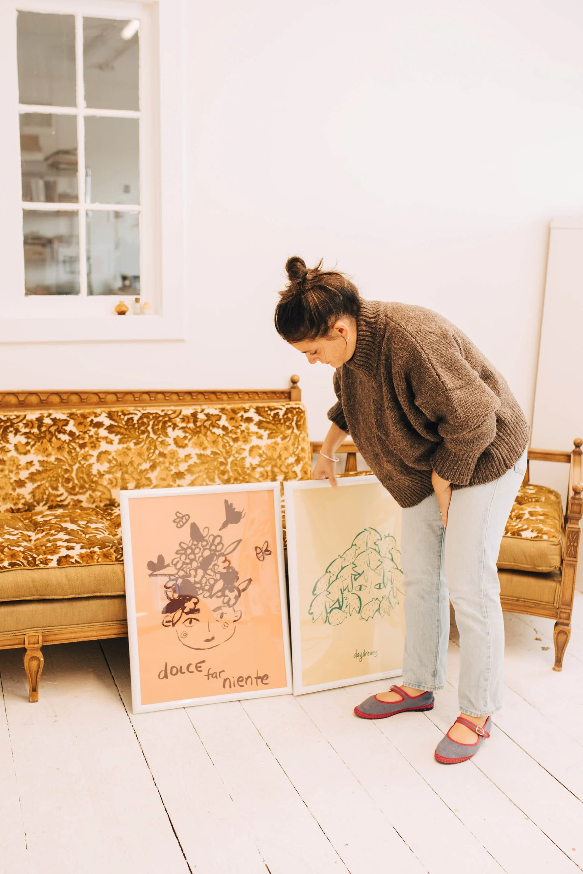 A woman looking down at artwork framed posters on a white wooden floor beside a patterned vintage sofa. The room has a white wall and window with reflection of a kitchen.