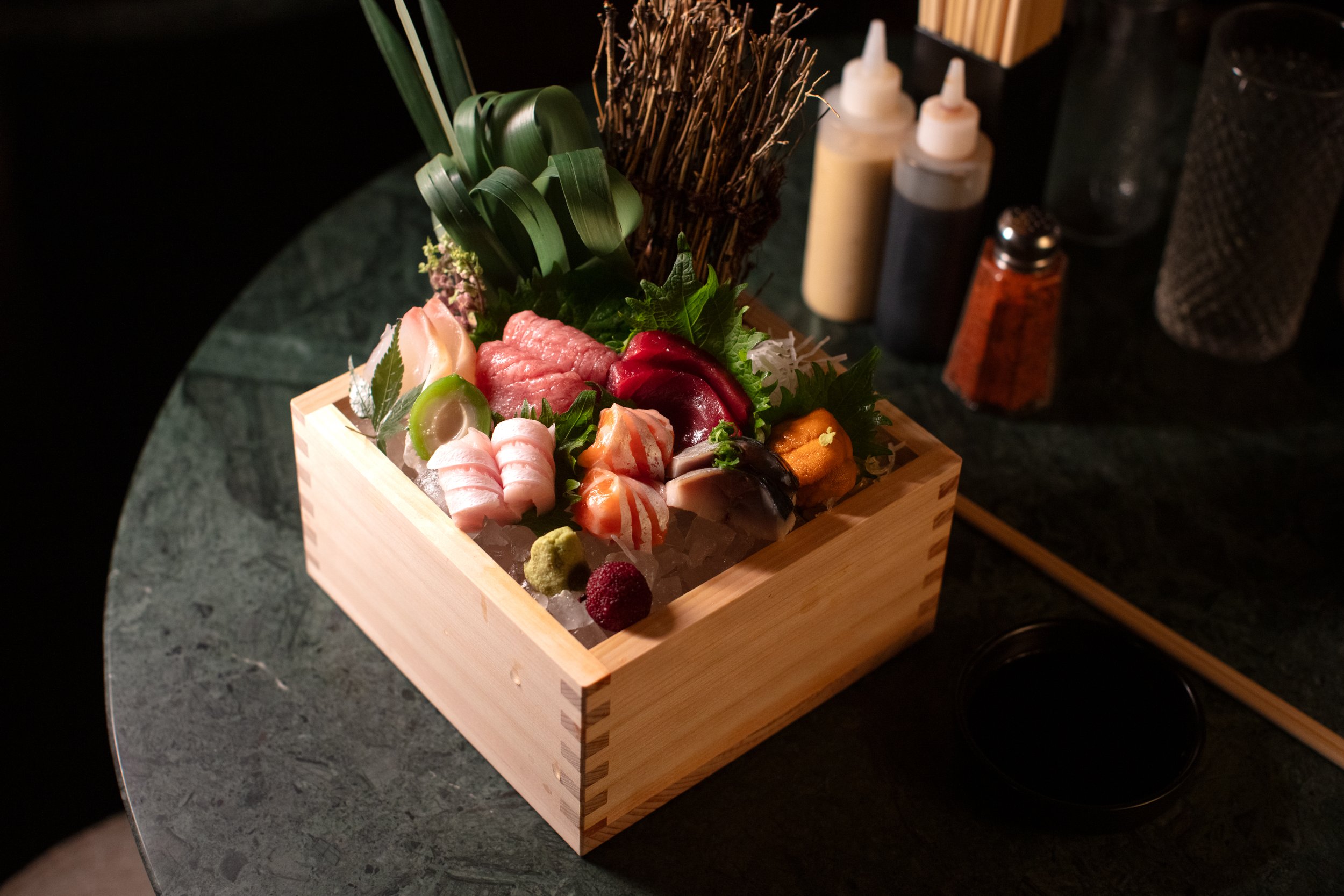Assorted sashimi platter in a wooden box on a table with soy sauce and condiments.