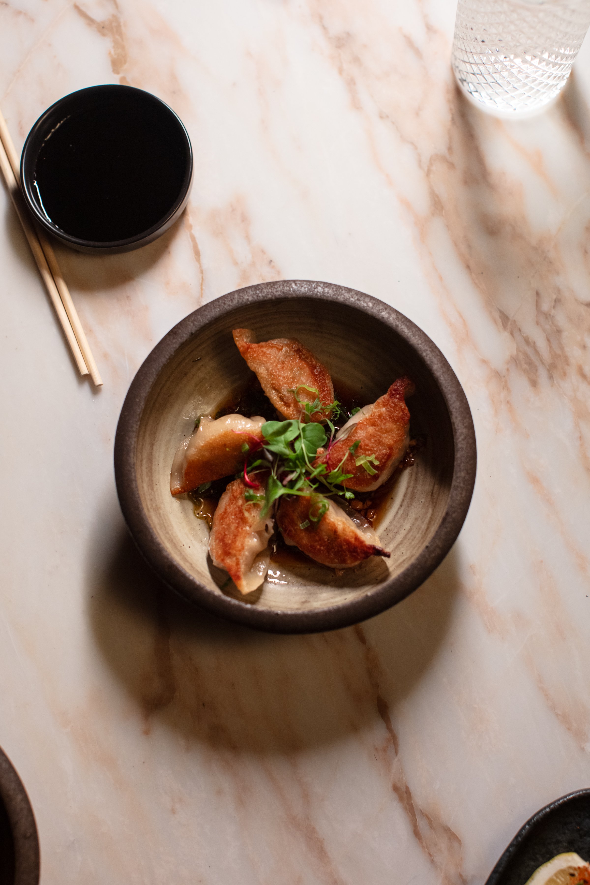 Bowl of pan-fried dumplings with greens, soy sauce dish, chopsticks, and glass on marble table.