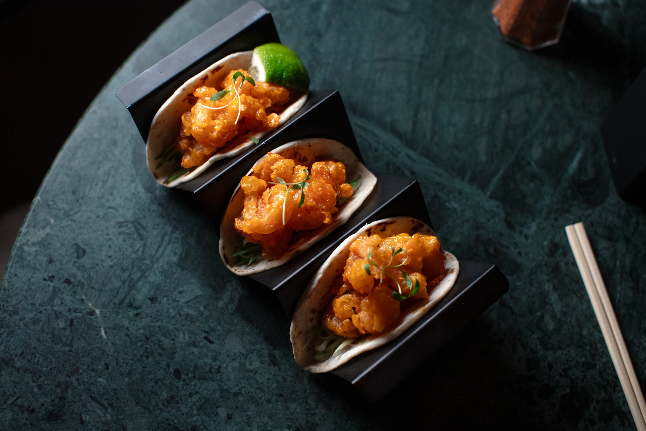 Three shrimp tacos in taco holders on a green marble table, garnished with herbs and a lime wedge.