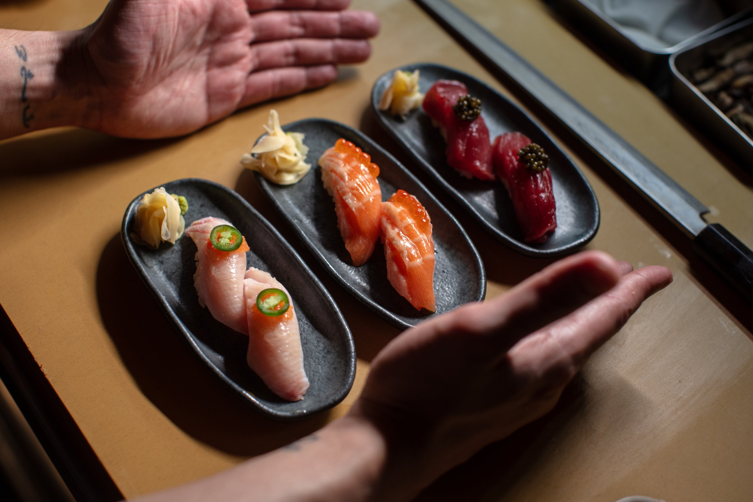 Assorted nigiri sushi on black plates with hands, knife, and garnishes