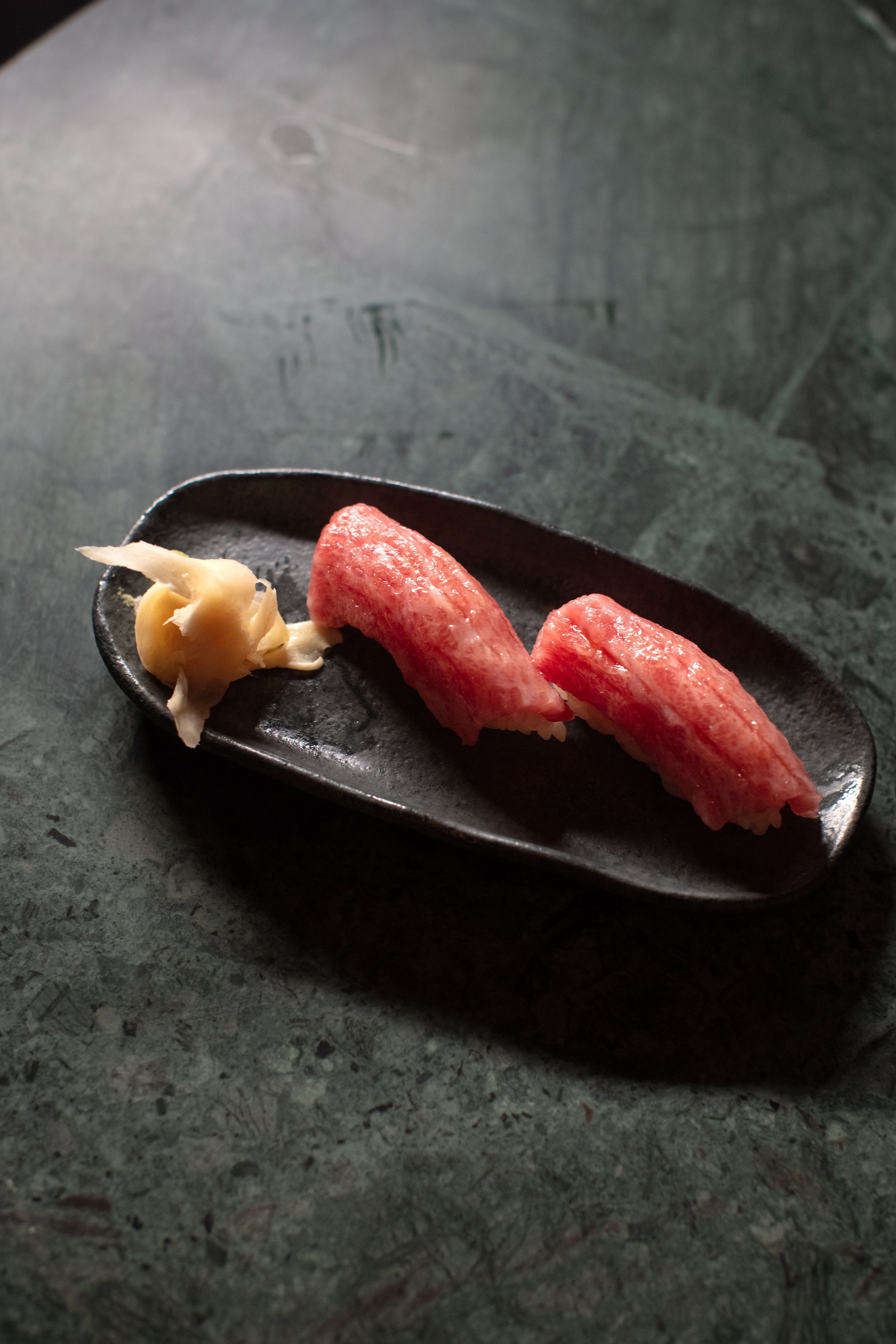 Two pieces of tuna nigiri sushi on a black plate with pickled ginger.
