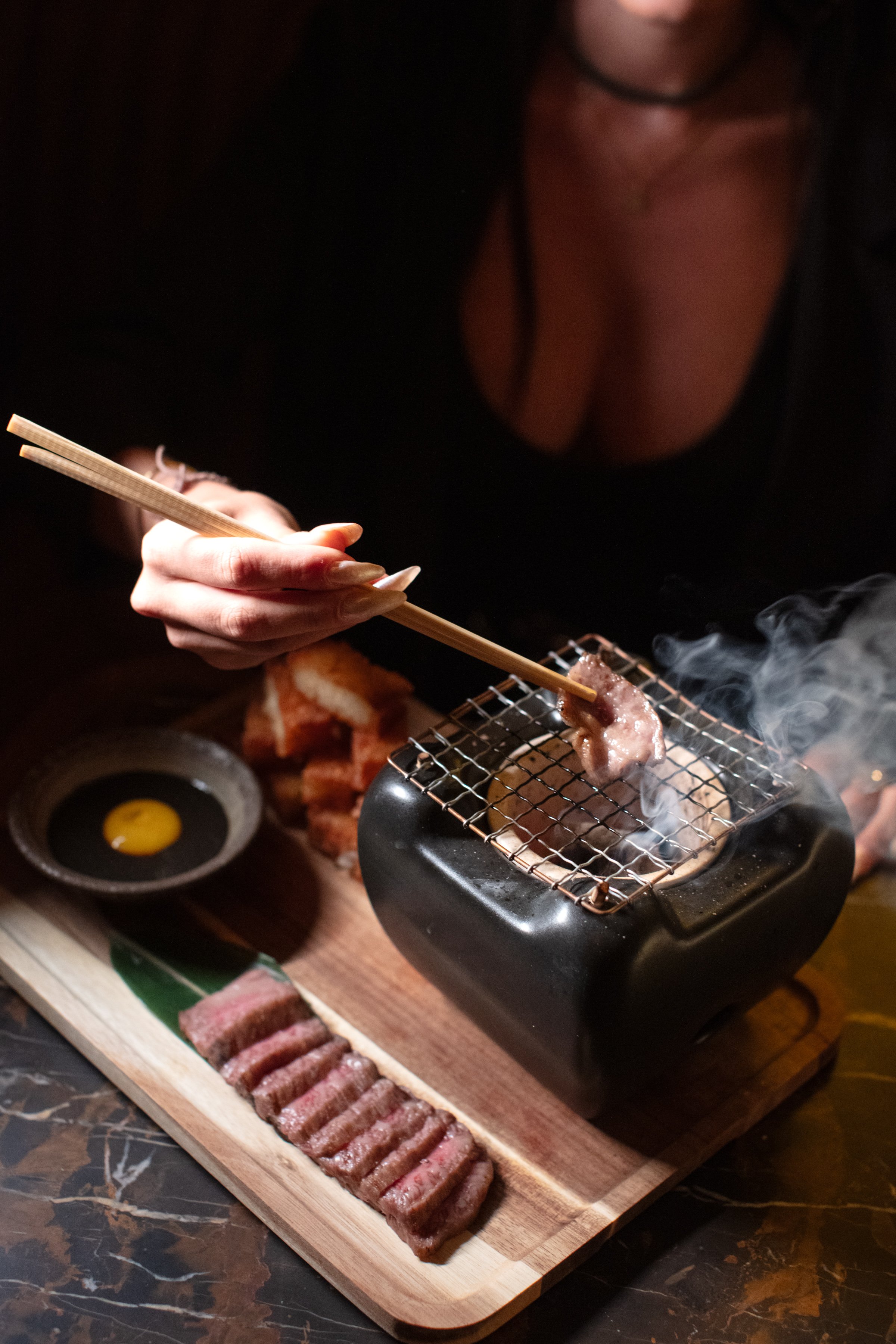Person grilling sliced meat over a small charcoal grill with chopsticks, accompanied by a dipping sauce and plated grilled meat on a wooden board.