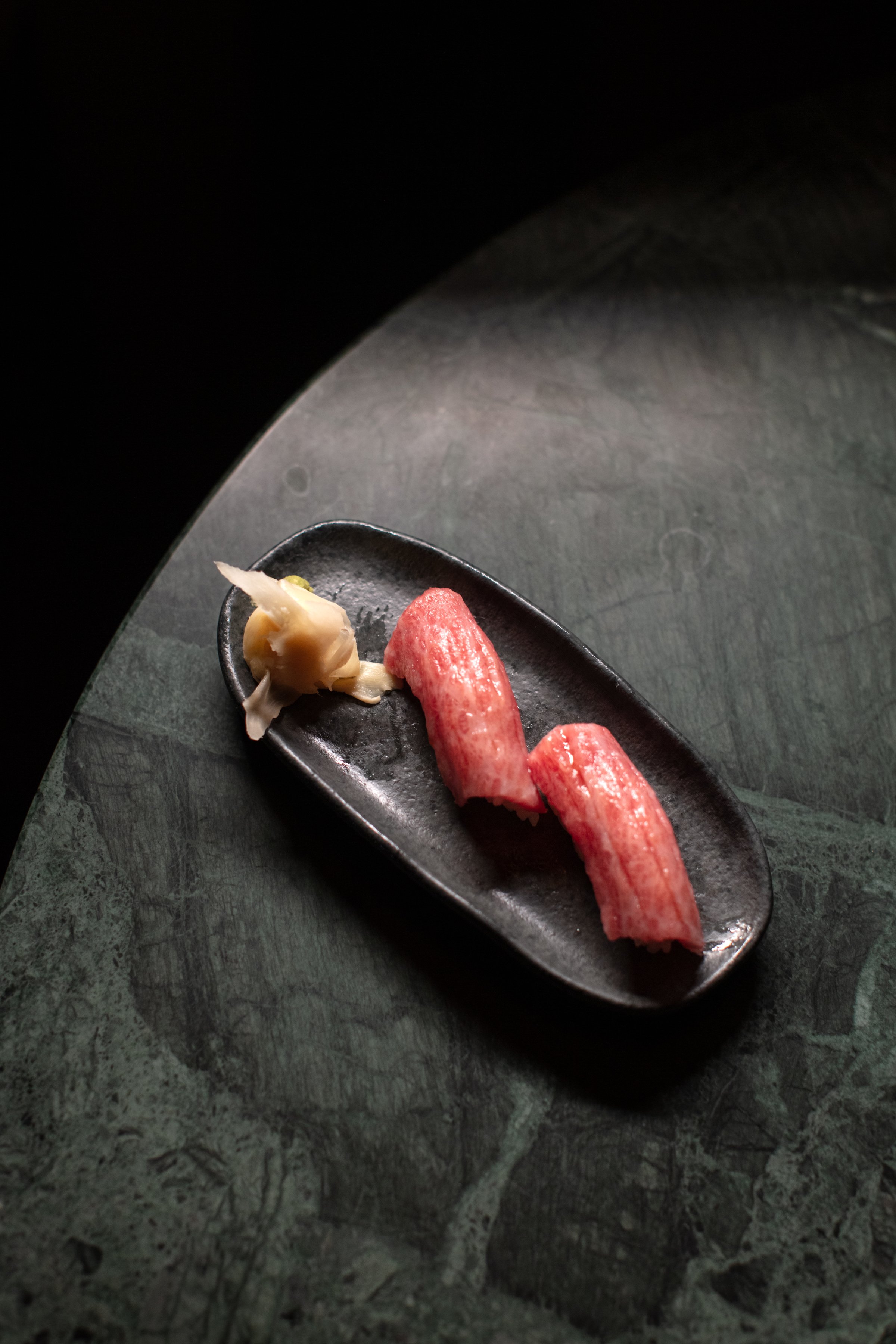Two pieces of sushi with pickled ginger on a black plate on a green marble surface.
