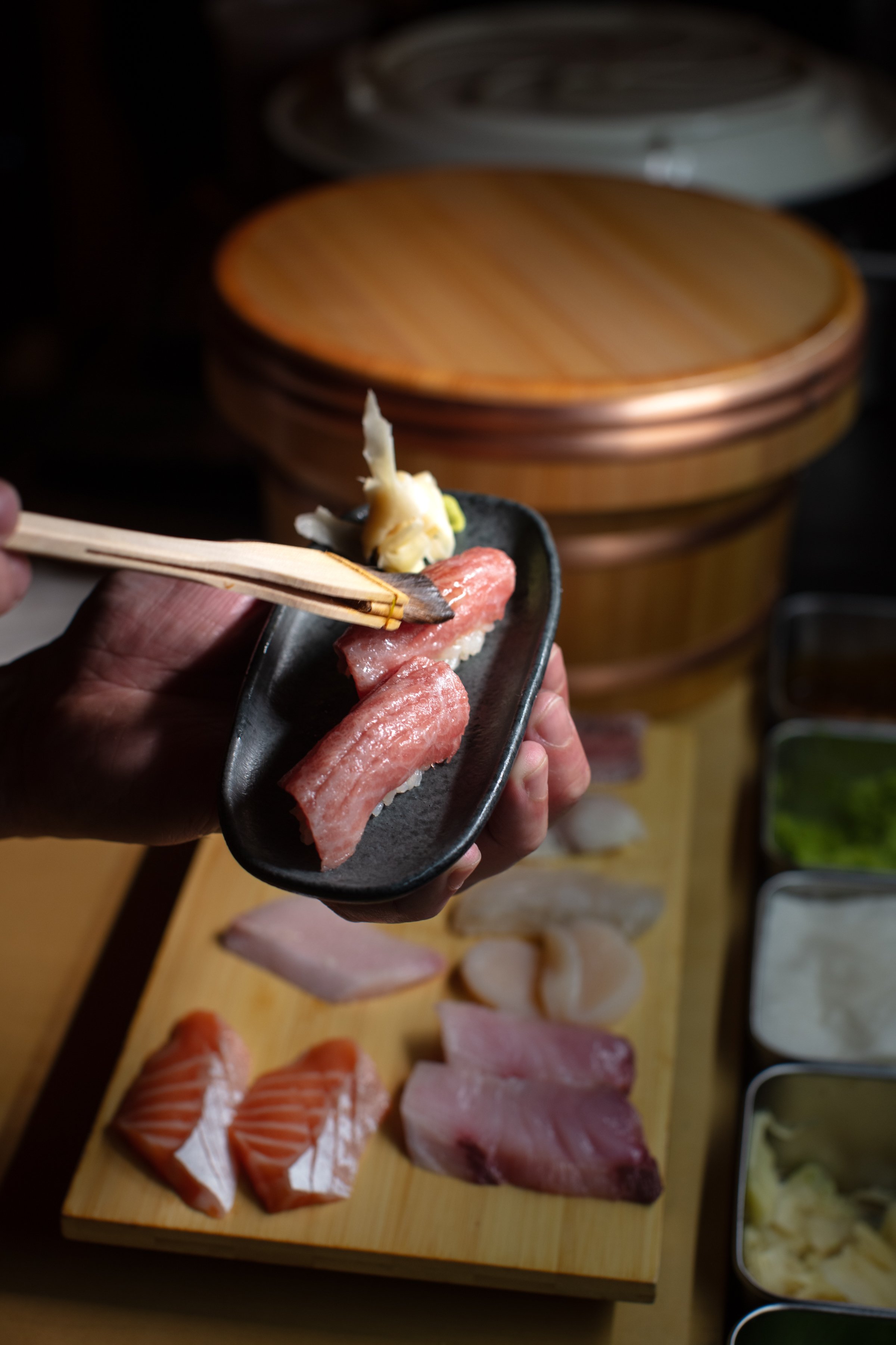Sushi presentation with various fish cuts on a wooden board; hand holding a dish with two pieces of sushi, garnished with wasabi and pickled ginger.