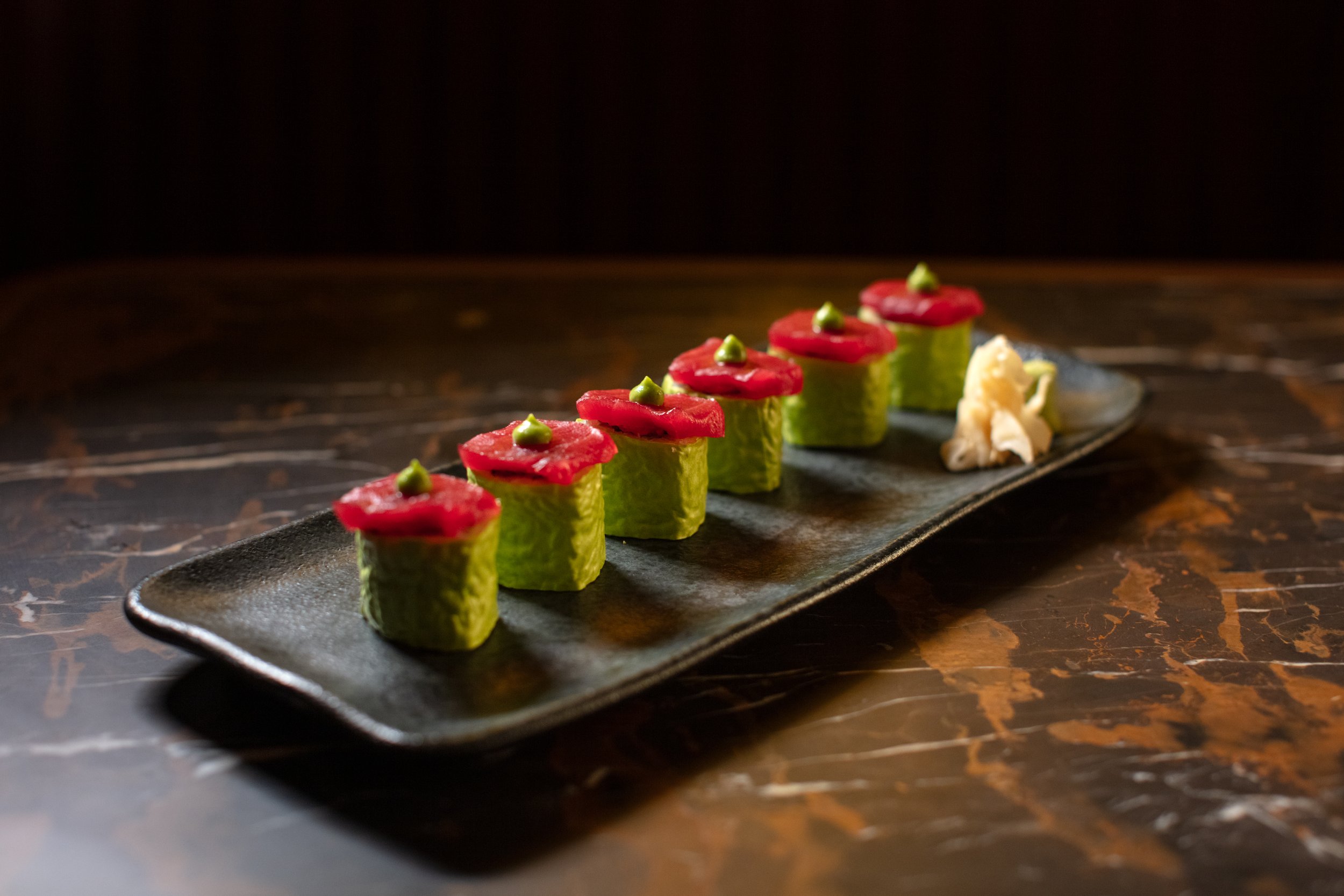 Sushi rolls wrapped in green vegetable leaves, topped with red fish and a dollop of wasabi, on a black plate with a side of pickled ginger.