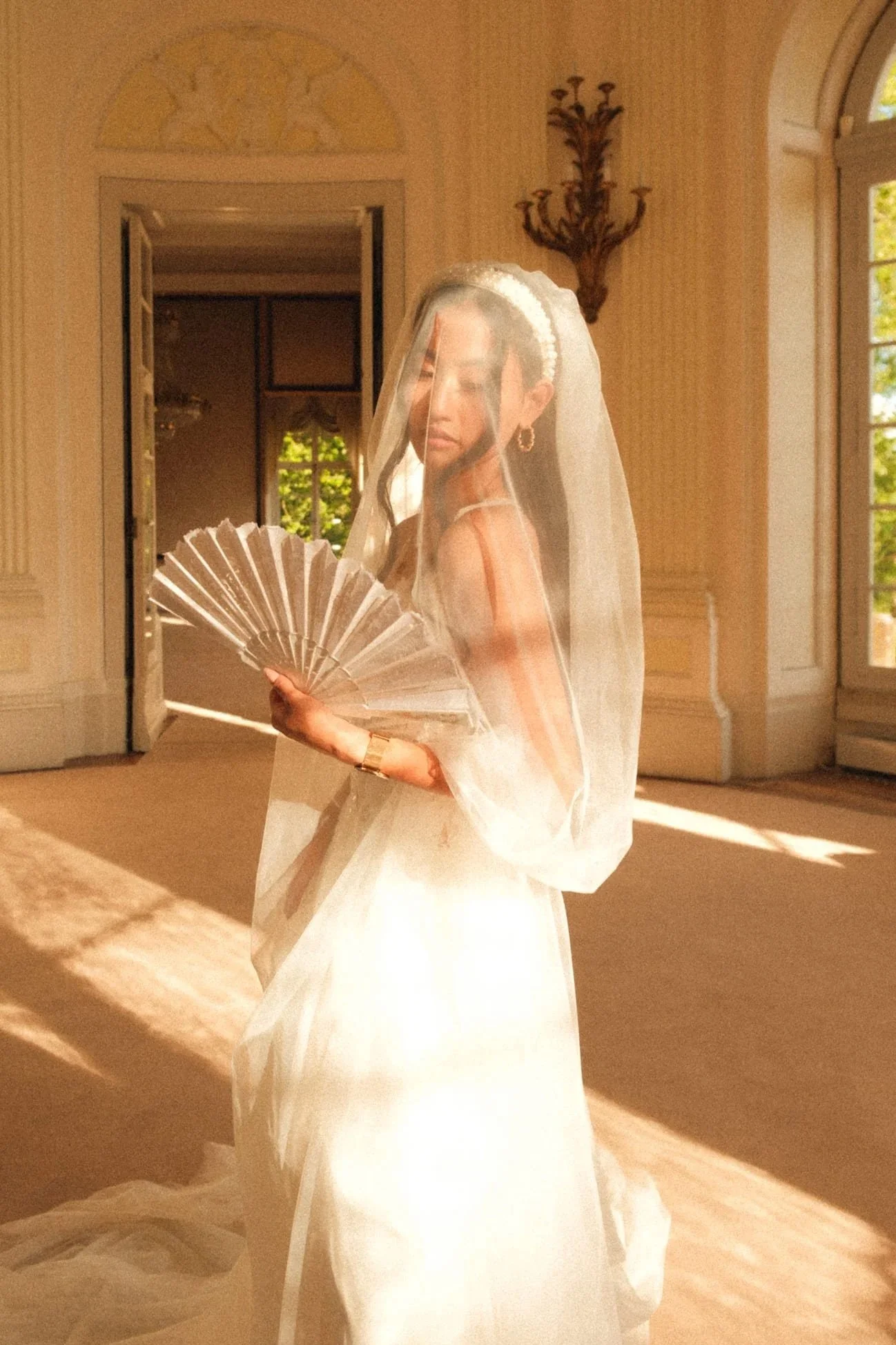 Frau in einem weißen Kleid mit Schleier und Diadem, hält einen Fächer, im Raum mit großen Fenstern und goldenen Wänden, Sonnenlicht strömt herein.