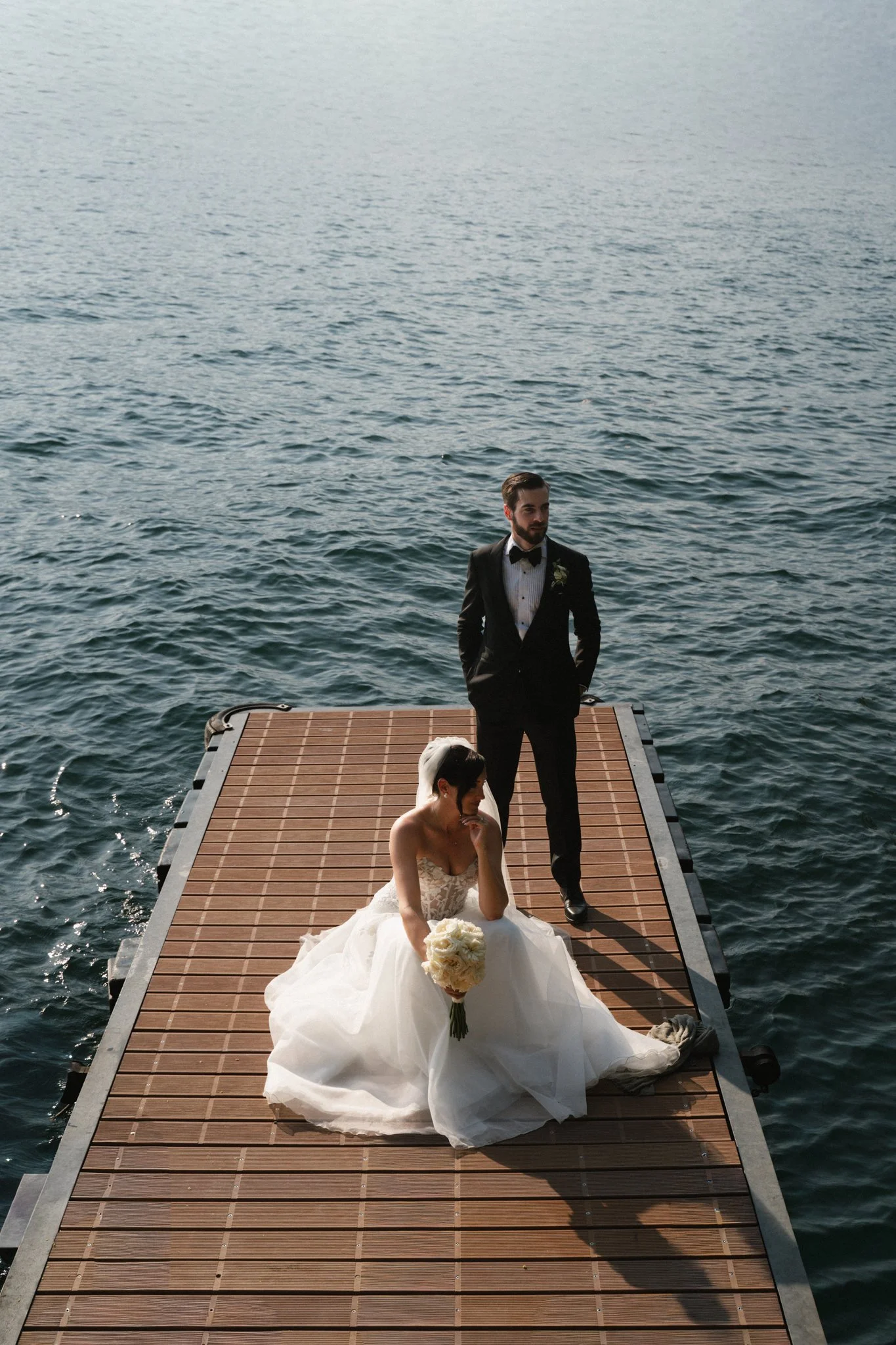 Braut und Bräutigam stehen auf einem Steg auf dem Wasser, die Braut sitzt im weißen Kleid mit Blumenstrauß, der Bräutigam im schwarzen Anzug und Fliege, der Himmel ist bewölkt.