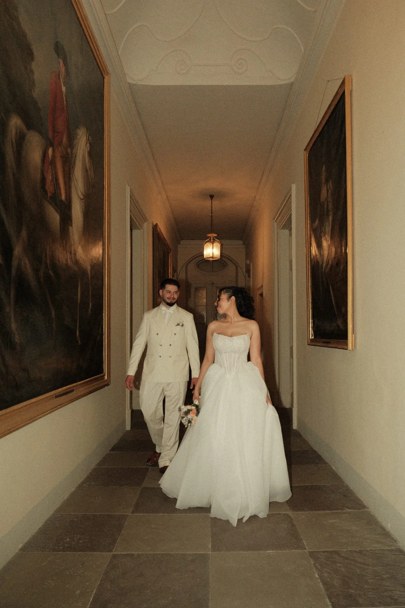 Ein Brautpaar geht Hand in Hand durch einen Flur in einem eleganten Gebäude mit Wandbildern und einer Pendelleuchte, die den Raum beleuchtet.