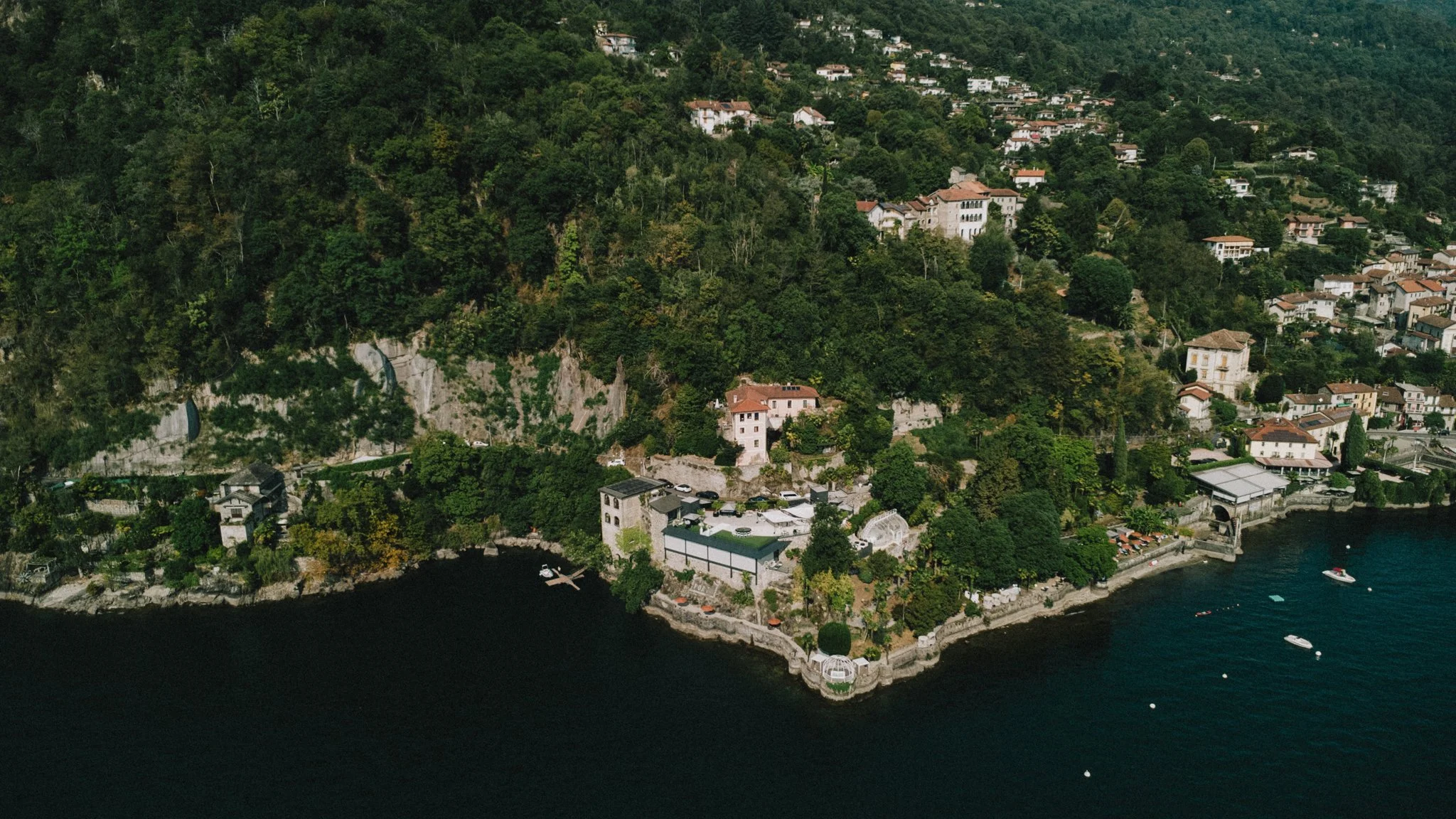 Ein Häuseransammlung an einer felsigen Küste mit Wasser im Vordergrund, umgeben von viel grünem Wald auf hügeligem Gelände.