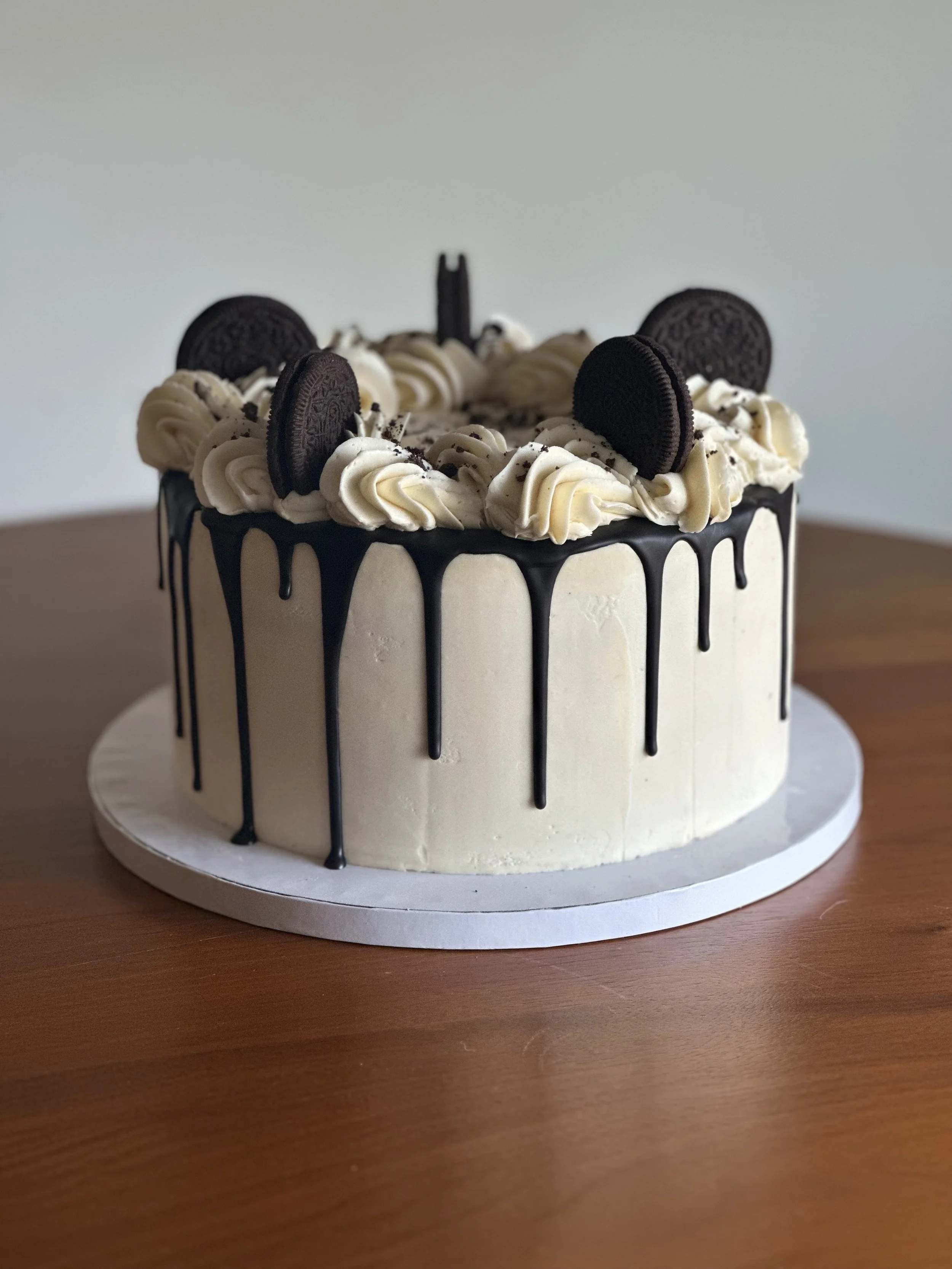 8" single-tier cake with vanilla buttercream frosting, chocolate drip, elaborate top piped border, and Oreo cookie crumbs and whole Oreo cookies on top.