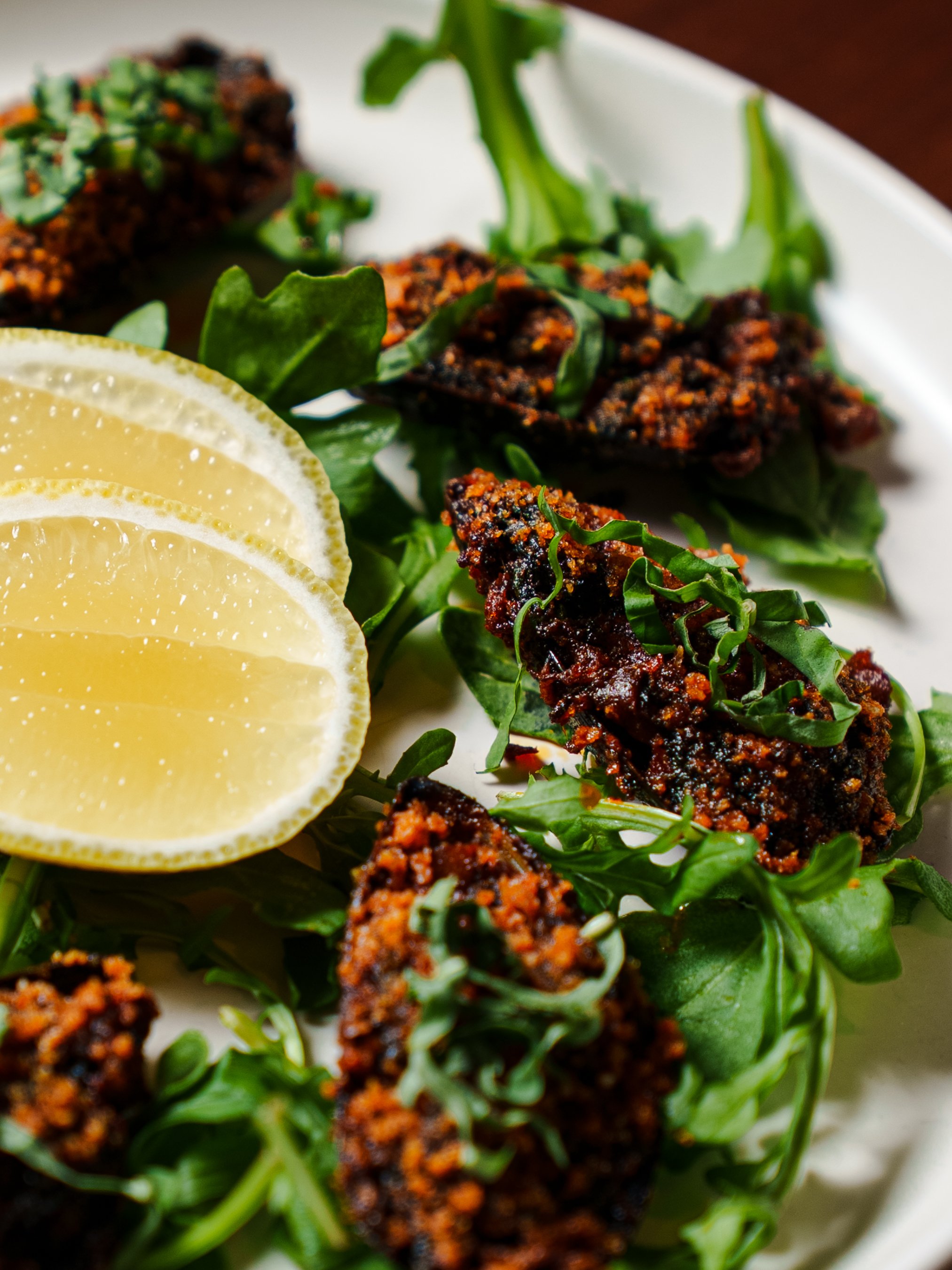 Fried chicken pieces with a crispy, dark coating, garnished with chopped herbs, served on a bed of leafy greens with lemon slices on the side.