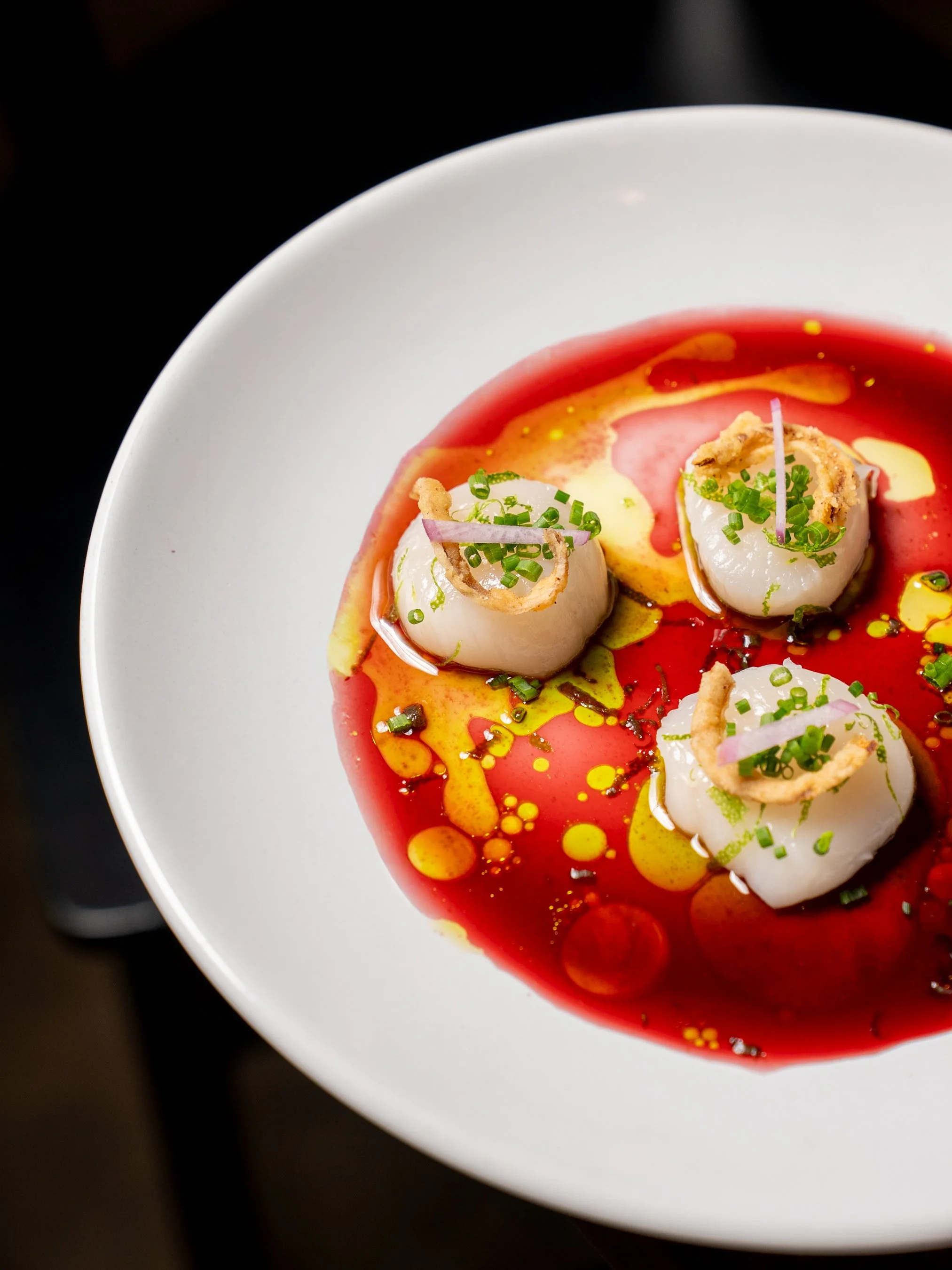 A white bowl containing three scallops served with a colorful sauce of yellow, red, and orange oils, garnished with chopped chives and thin onion slices.