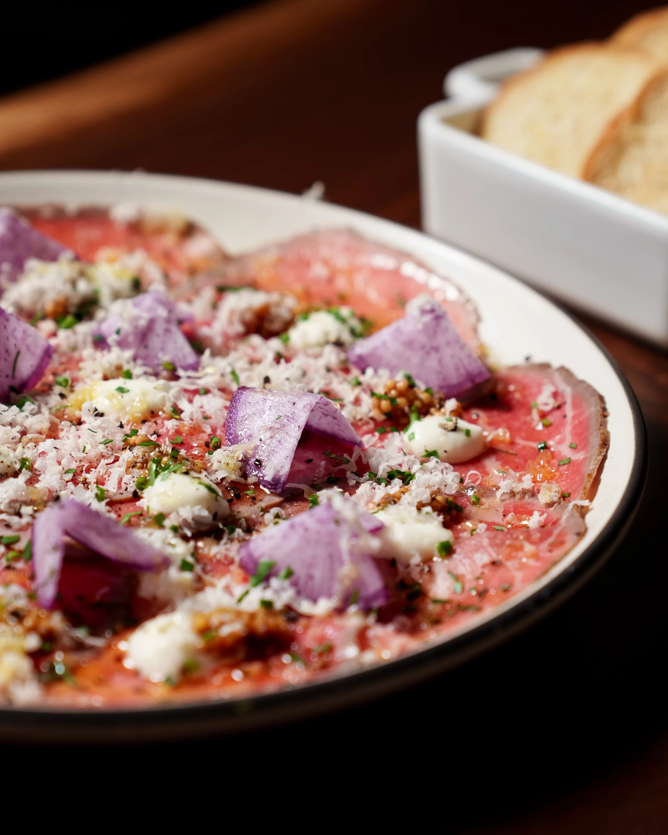 Thinly sliced raw beef carpaccio topped with purple onion pieces, grated cheese, chopped herbs, and capers, served with slices of bread in the background.