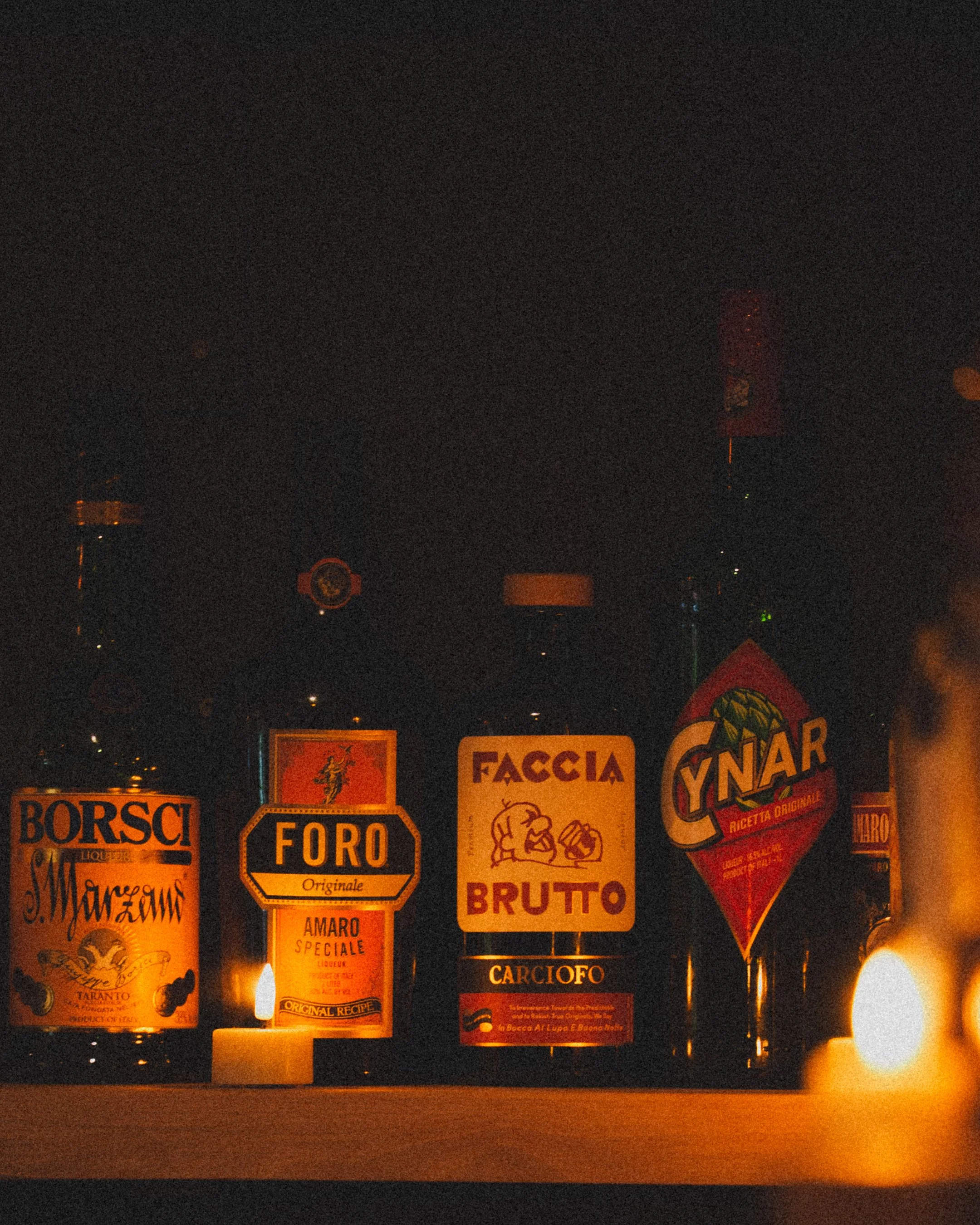 A row of various bottles with Italian-style labels illuminated by candlelight in a dark setting.