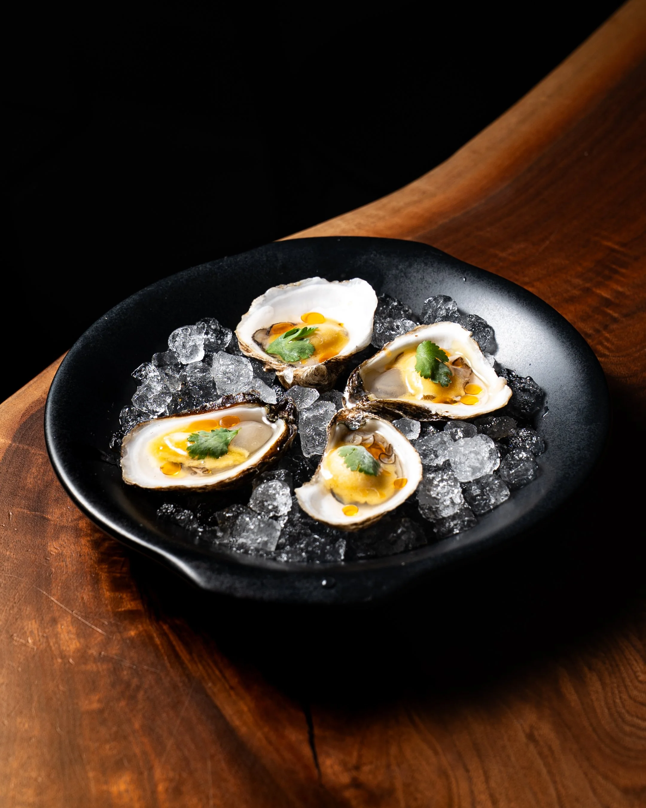 Four oysters on a bed of ice in a black bowl, garnished with cilantro leaves, on a wooden surface.