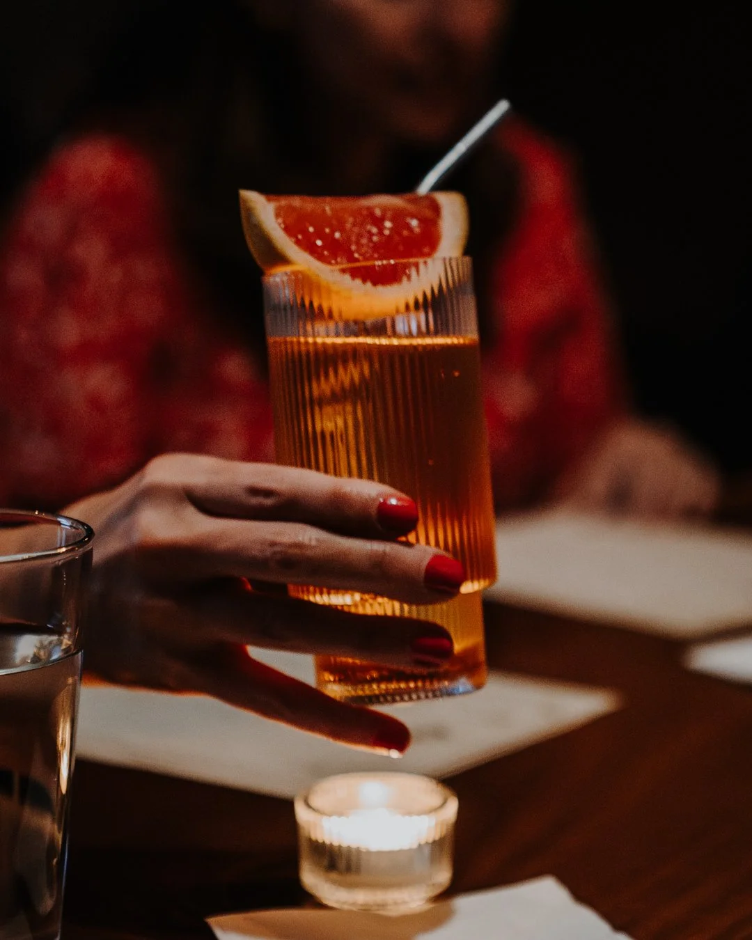Person holding a tall glass of brownish-orange drink with a wedge of grapefruit and a straw, seated at a table with a lit candle and a glass of water nearby.