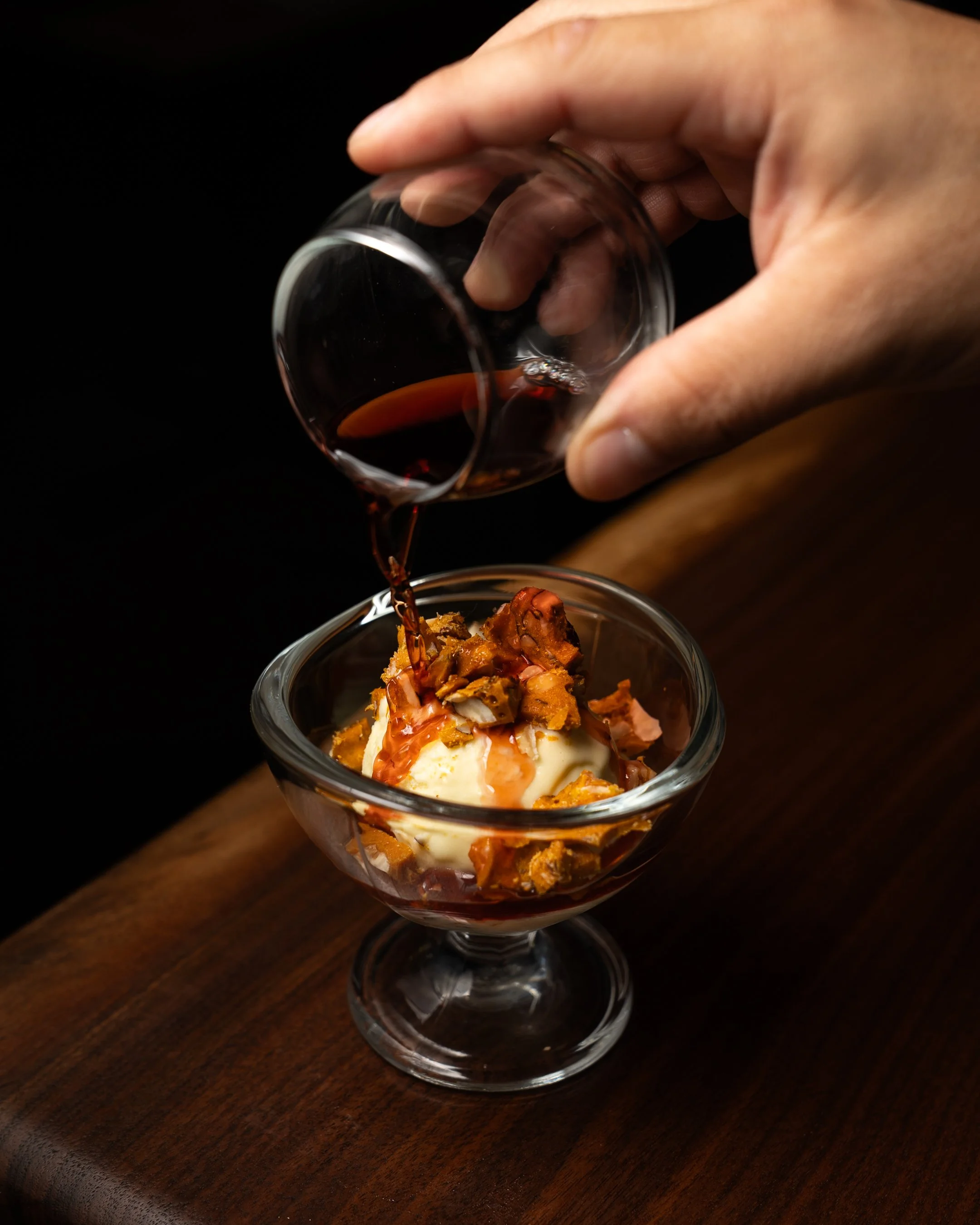 A hand pouring red wine over a dessert with what appears to be ice cream, caramelized fruit, and crispy topping in a glass cup on a wooden surface.