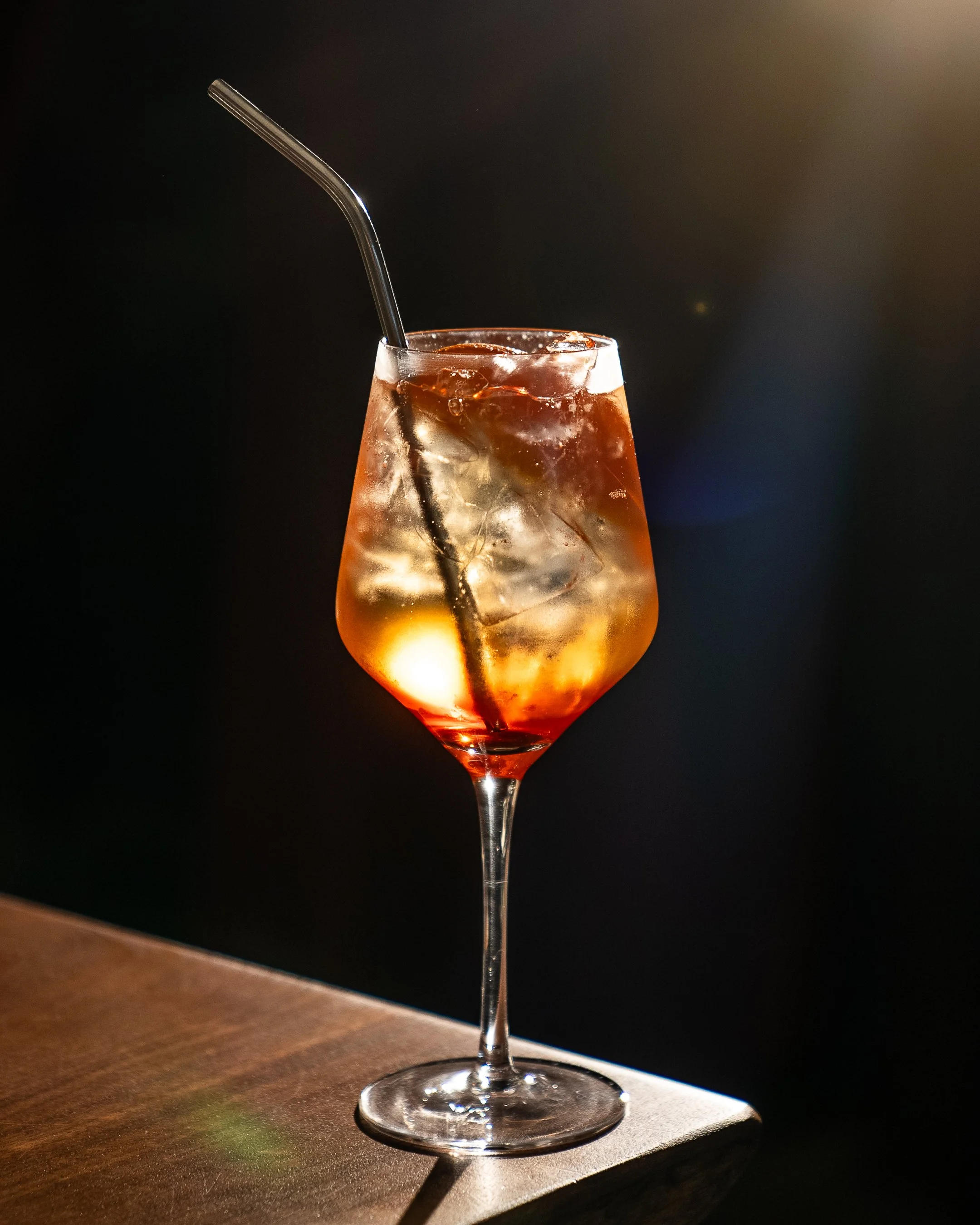 A tall wine glass filled with a dark, fizzy beverage with ice and a black straw, set on a wooden surface with a dark background and warm light glare.