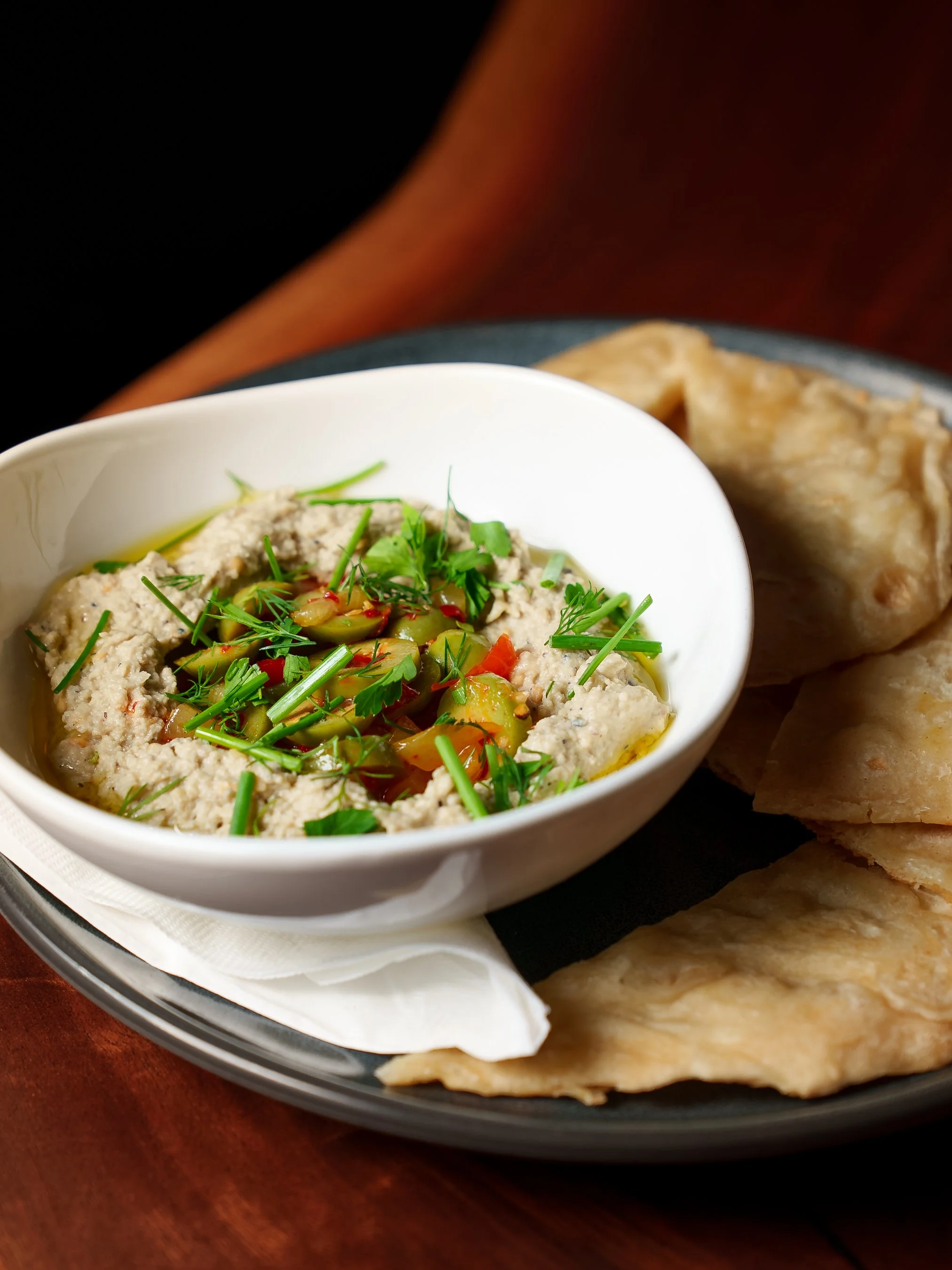 A white bowl filled with a creamy hummus topped with chopped herbs and cherry tomatoes, accompanied by pieces of flatbread on a dark plate.