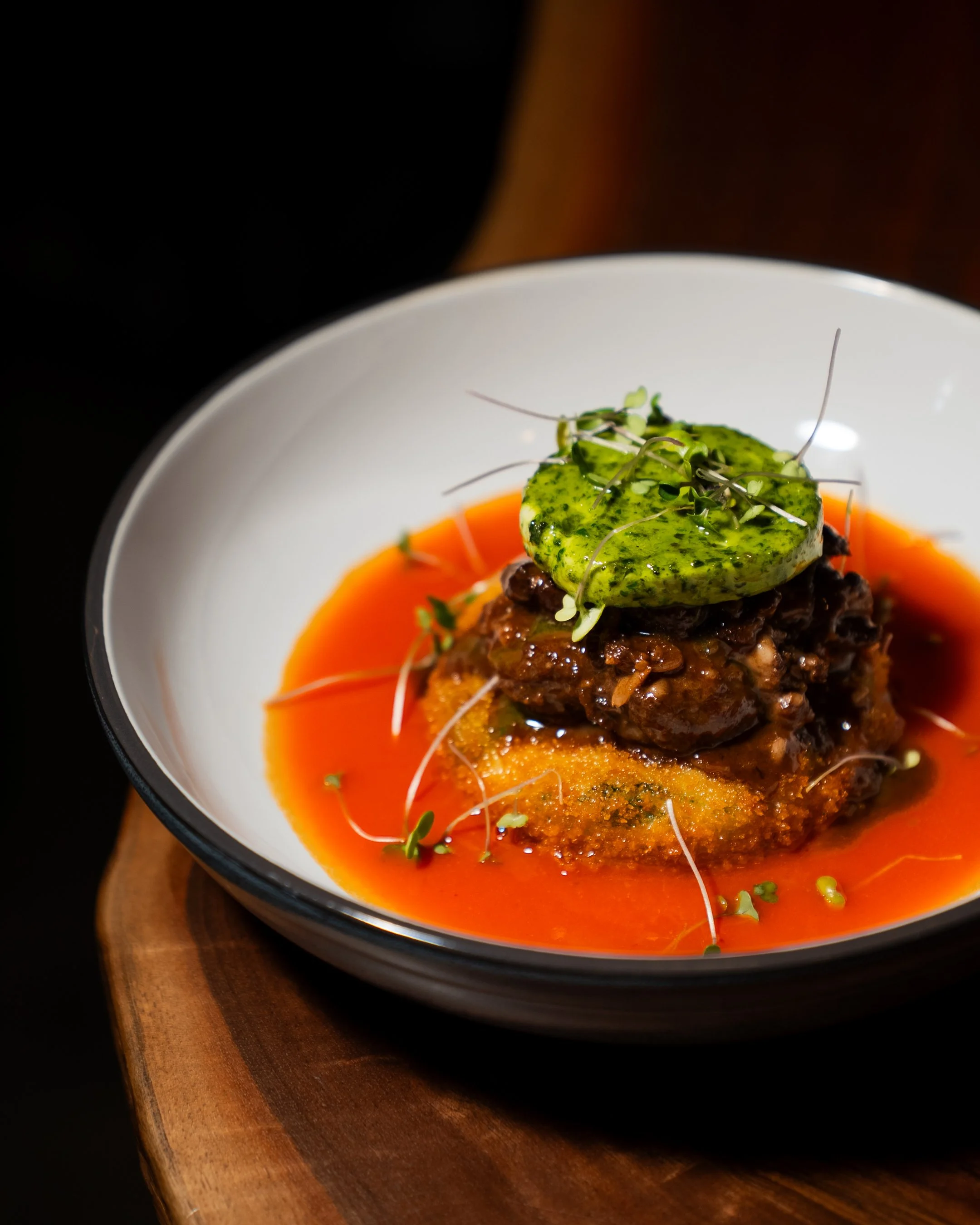 A plated gourmet dish featuring sauce, meat, and a green garnish, served on a white bowl placed on a wooden surface.