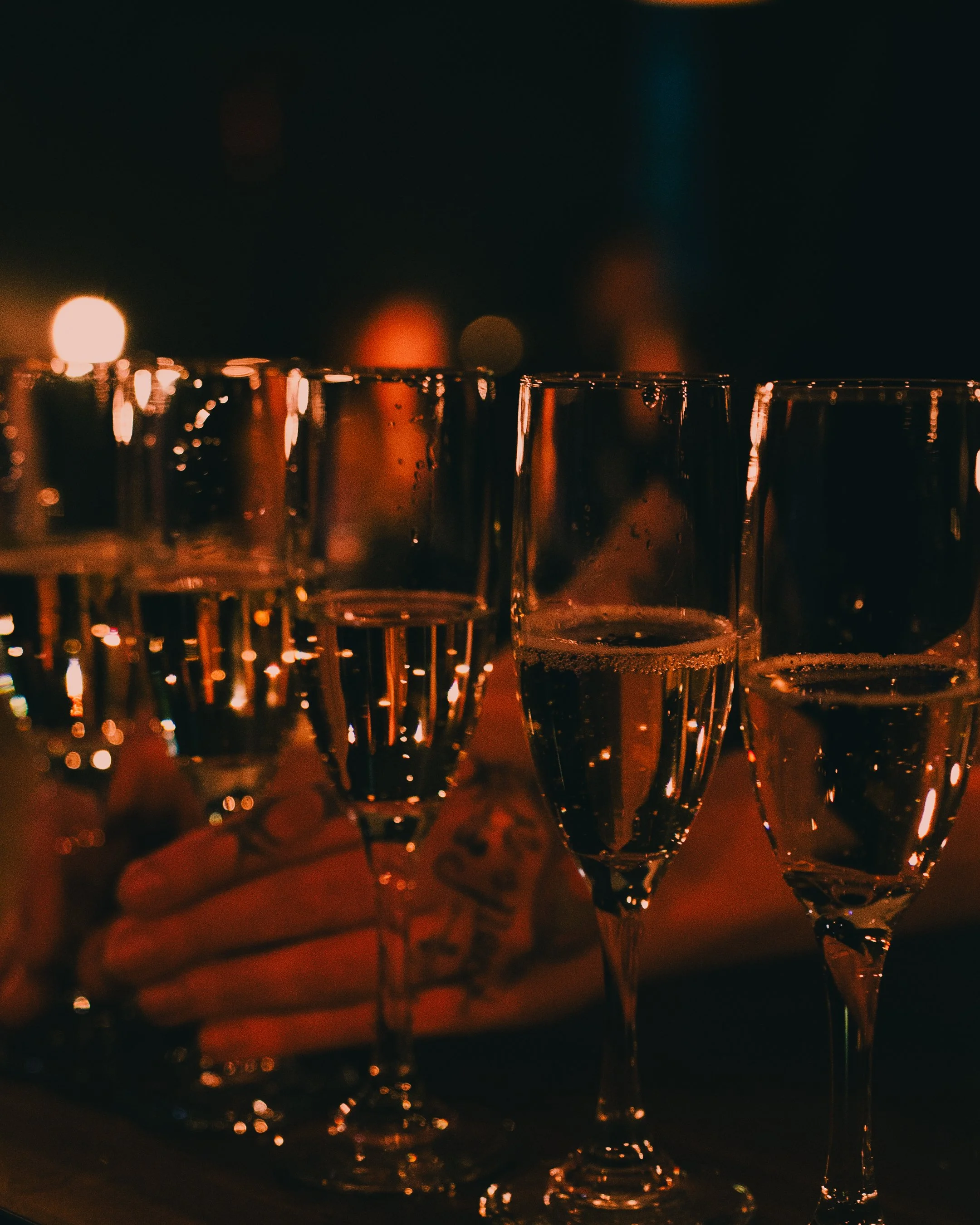 Several champagne flutes filled with champagne, held by a person's hand, against a dark background with bokeh lights.