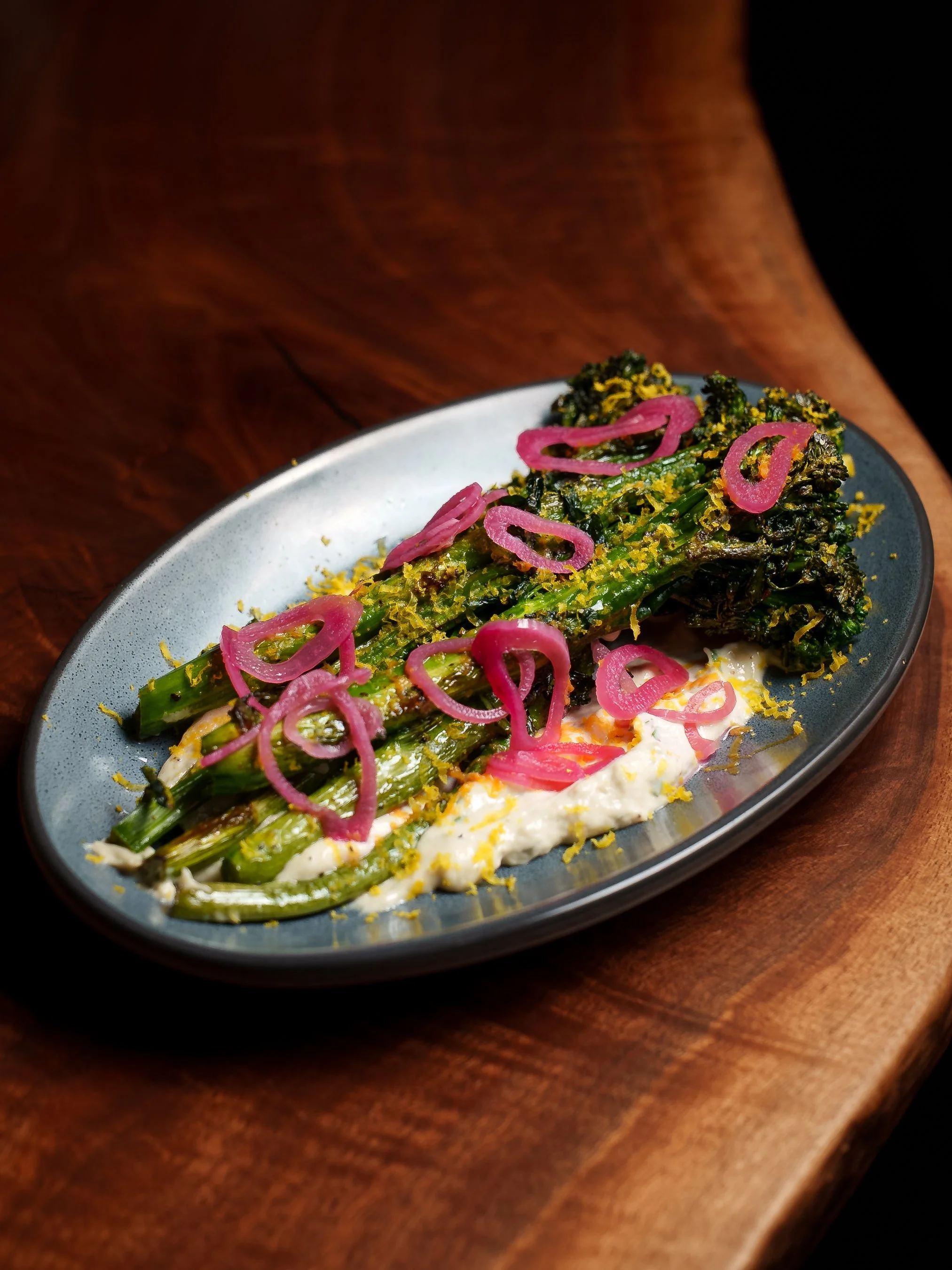 Grilled asparagus with pickled red onions, lemon zest, and a creamy sauce on a gray oval plate on a wooden surface.